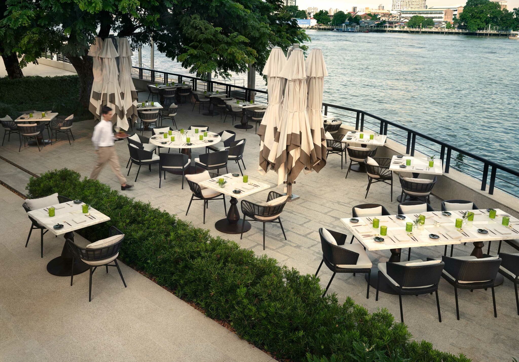 Riverside restaurant patio with empty tables set with green glasses and white napkins, surrounded by greenery and large umbrellas near the water.