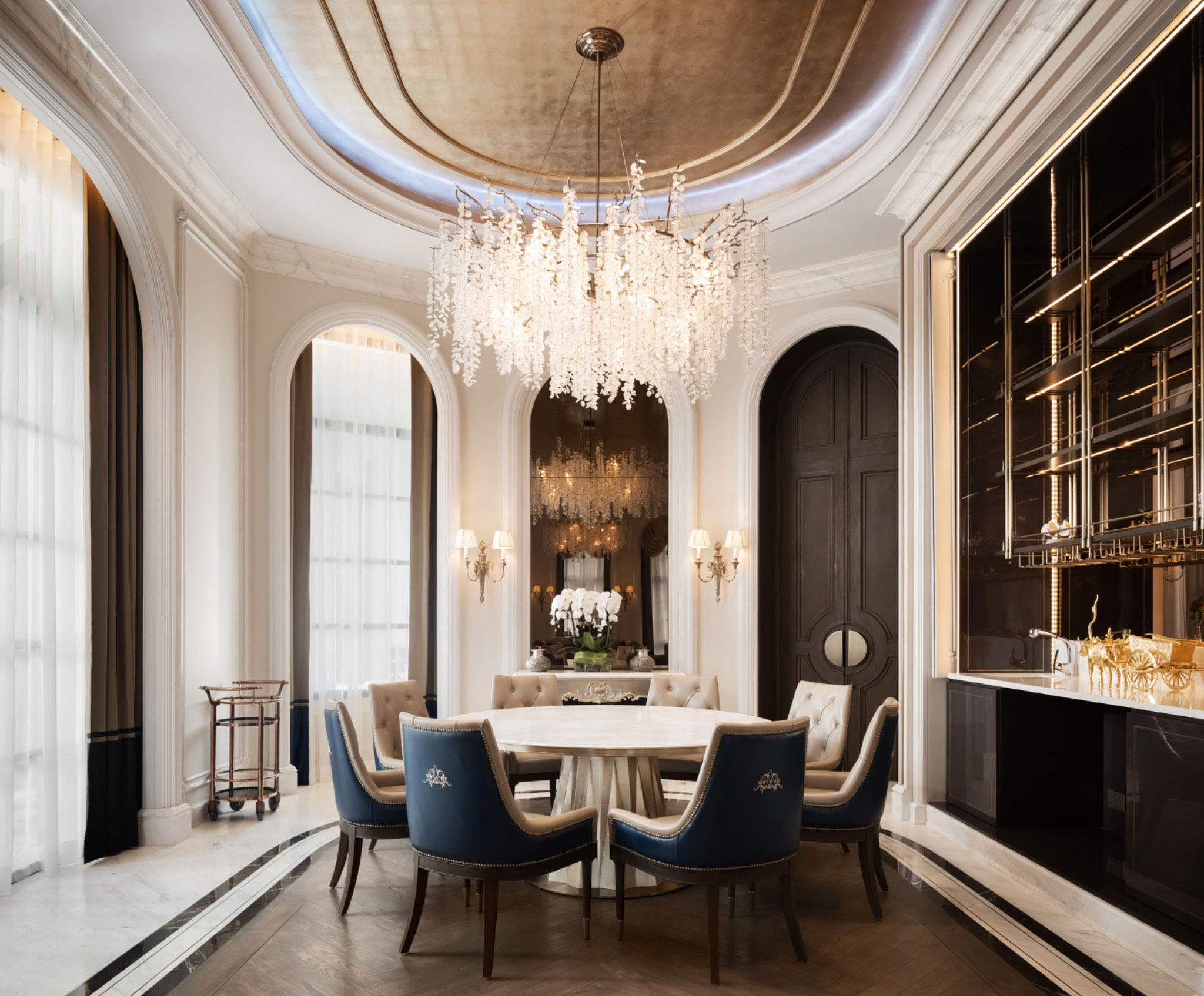 Elegant dining room with a round table, blue and beige chairs, ornate chandelier, and a large mirror. Luxurious and sophisticated ambiance.