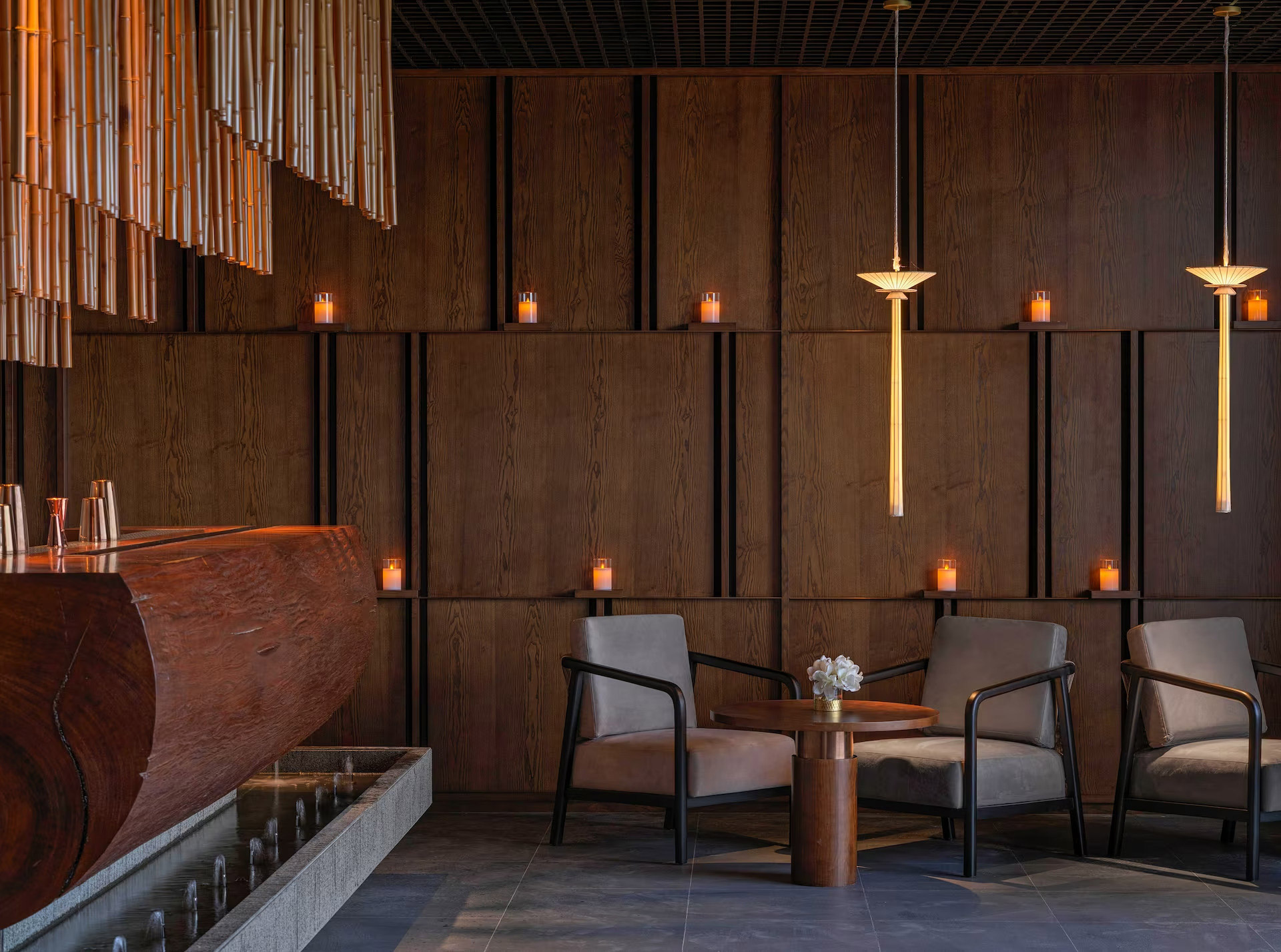 Sophisticated hotel restaurant with dark wood paneling, soft ambient lighting from candles, pendant lights, two cushioned chairs, and a small round table.