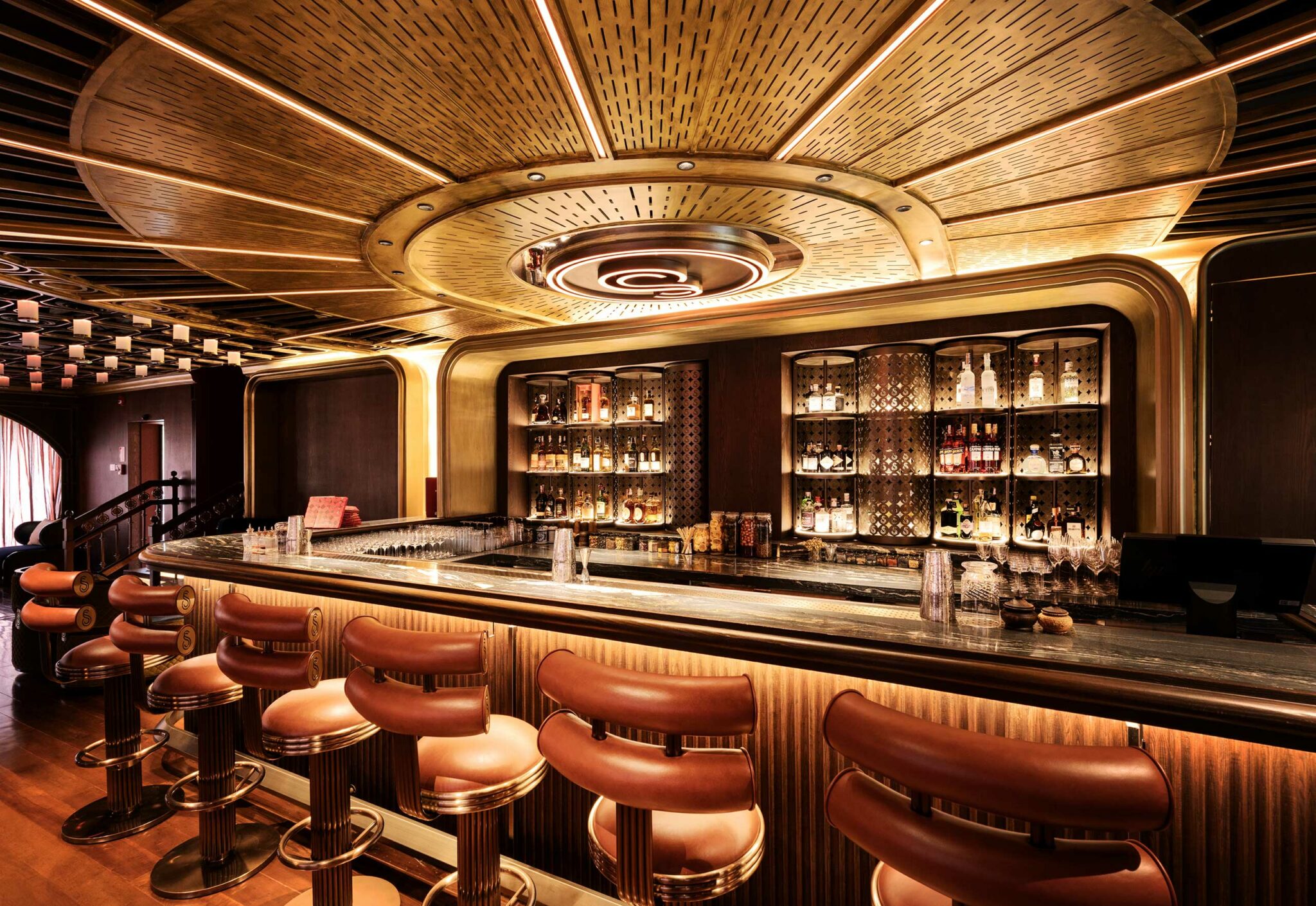 Luxurious restaurant bar area with warm lighting, featuring plush brown stools, a sleek marble counter, and a stocked liquor shelf. The gold ceiling adds elegance.