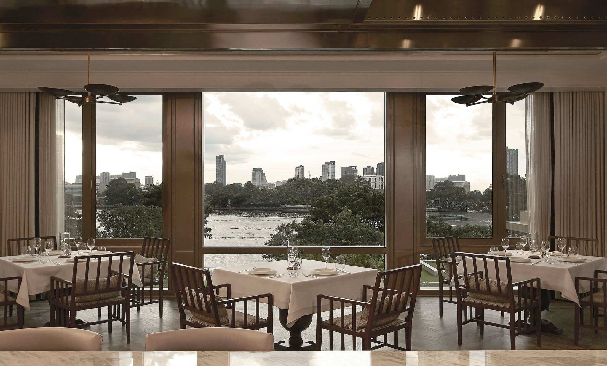 Elegant restaurant interior with wooden chairs and white-clothed tables, overlooking a river and city skyline through large windows. Calm and inviting ambiance.