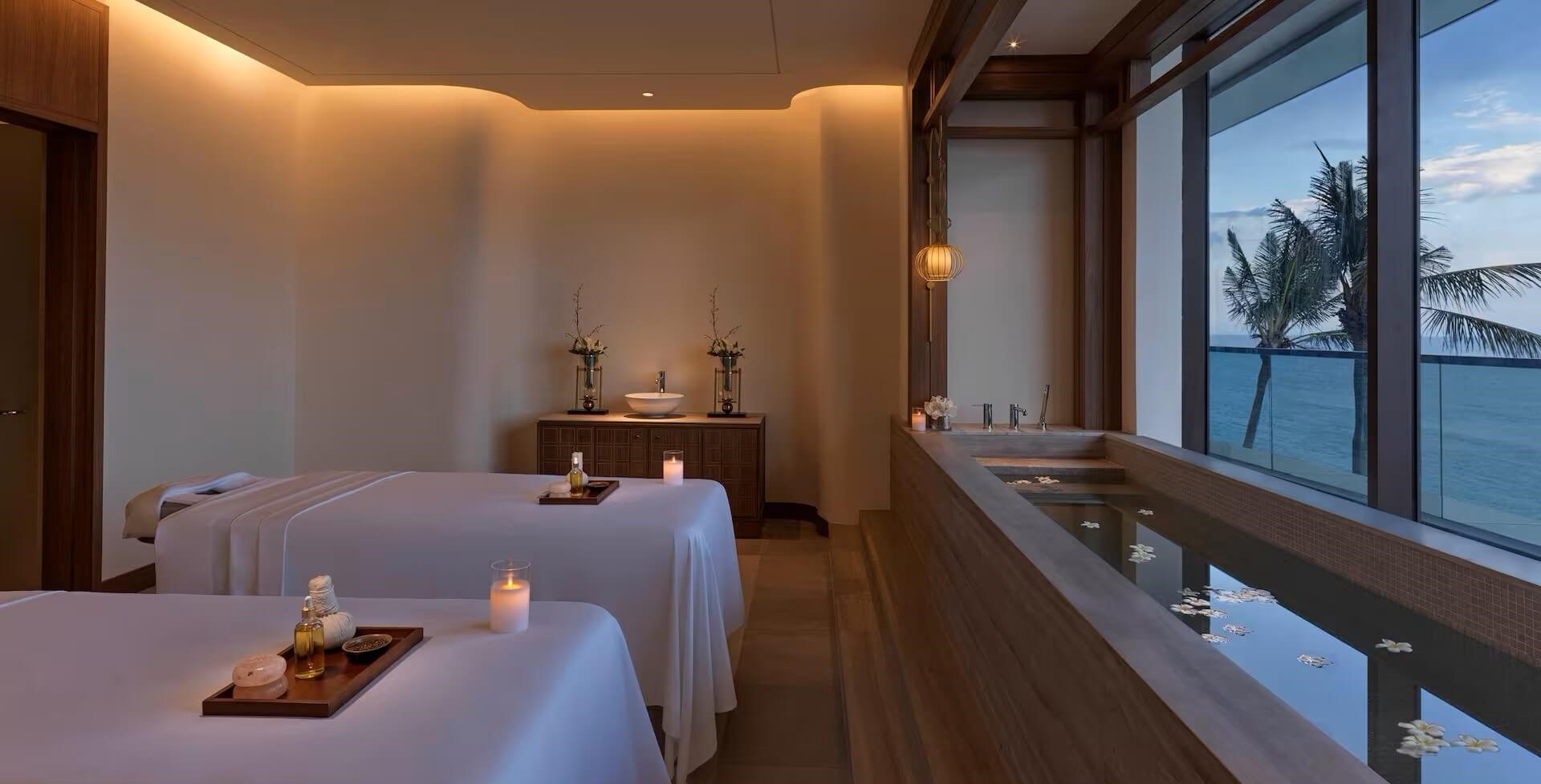 Serene hotel spa treatment room with two massage tables, soft candlelight, and ocean views through large windows. A wooden tub is filled with floating flowers, creating a tranquil atmosphere.