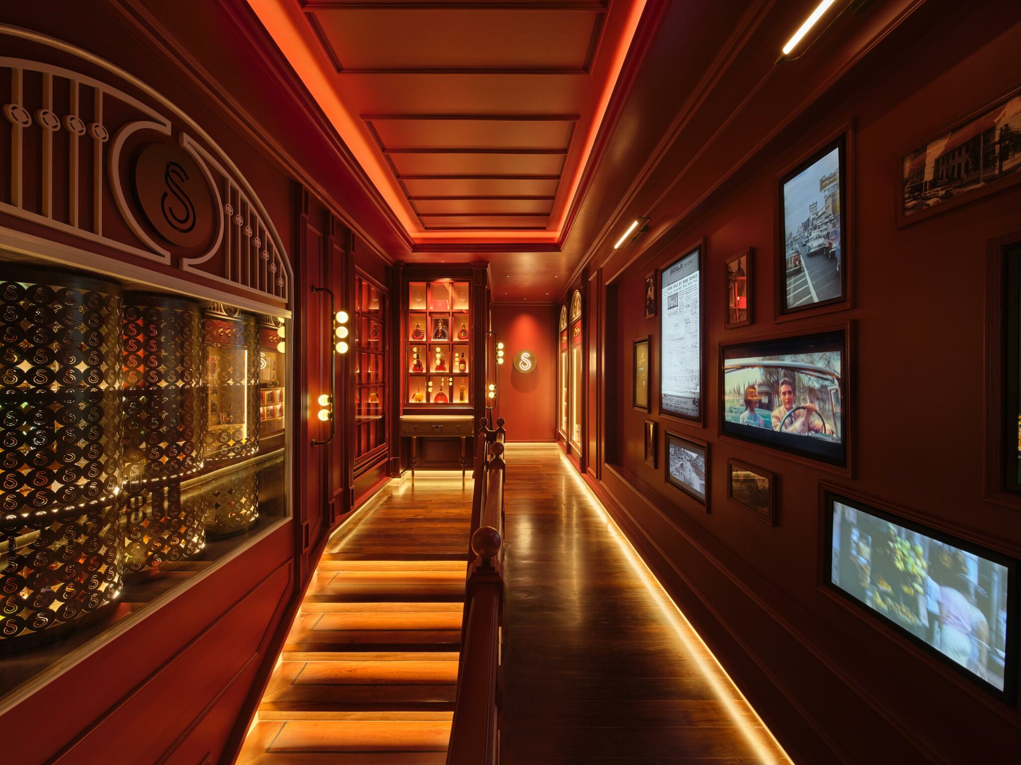 Dimly lit restaurant hallway with rich red walls and warm lighting. Shelves display bottles; framed photos adorn the right wall. Elegant and inviting atmosphere.