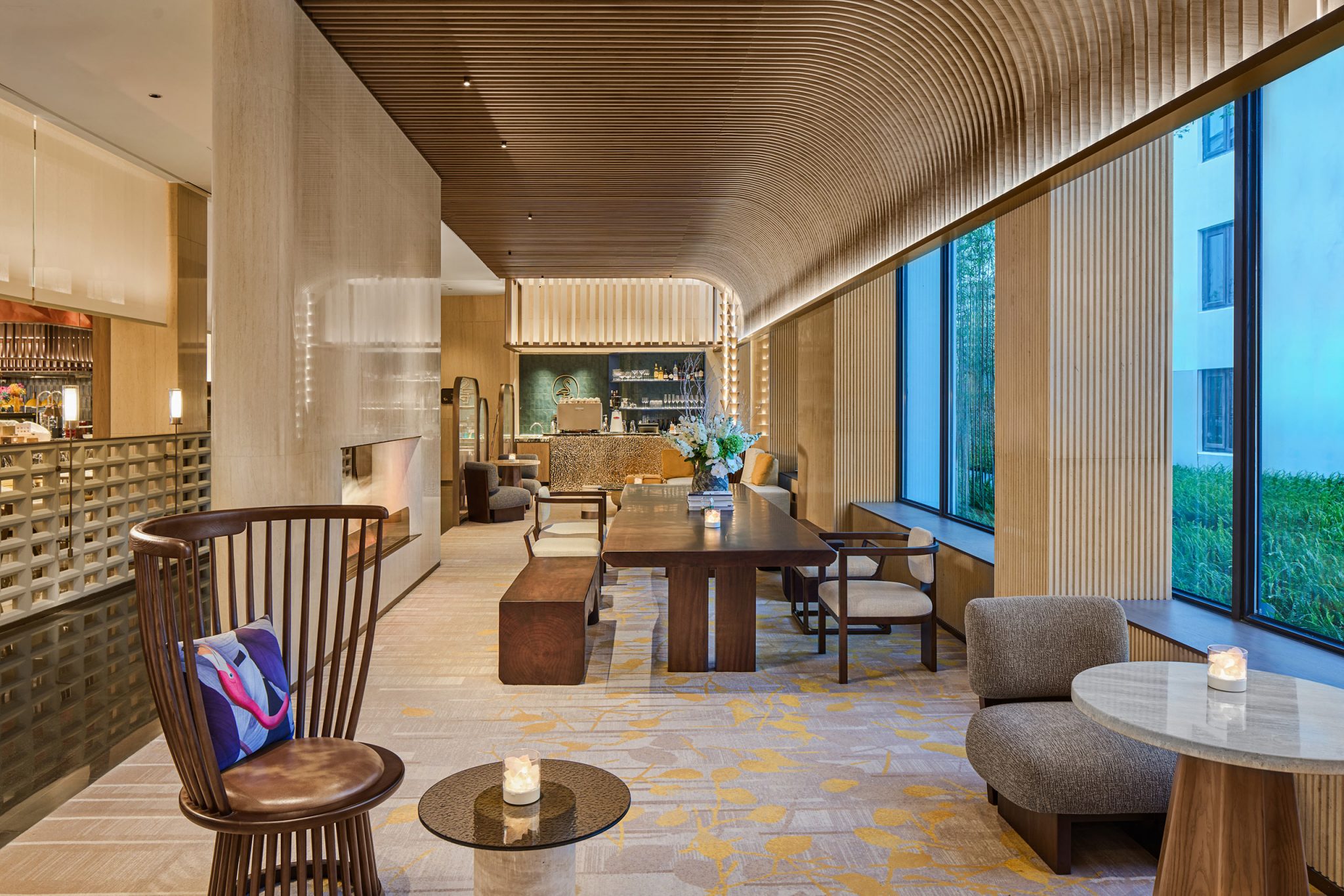 Modern, elegant hotel lounge with wooden ceiling and warm lighting. Features plush seating, a large table, decorative plants, and a bar in the background.