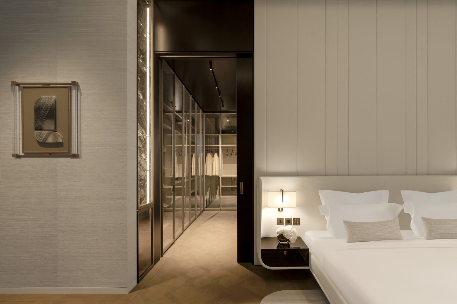 Modern luxurious bedroom with a neatly made bed, bedside table, illuminated lamp, wall art, and a view into a glass-walled walk-in closet with hanging clothes.