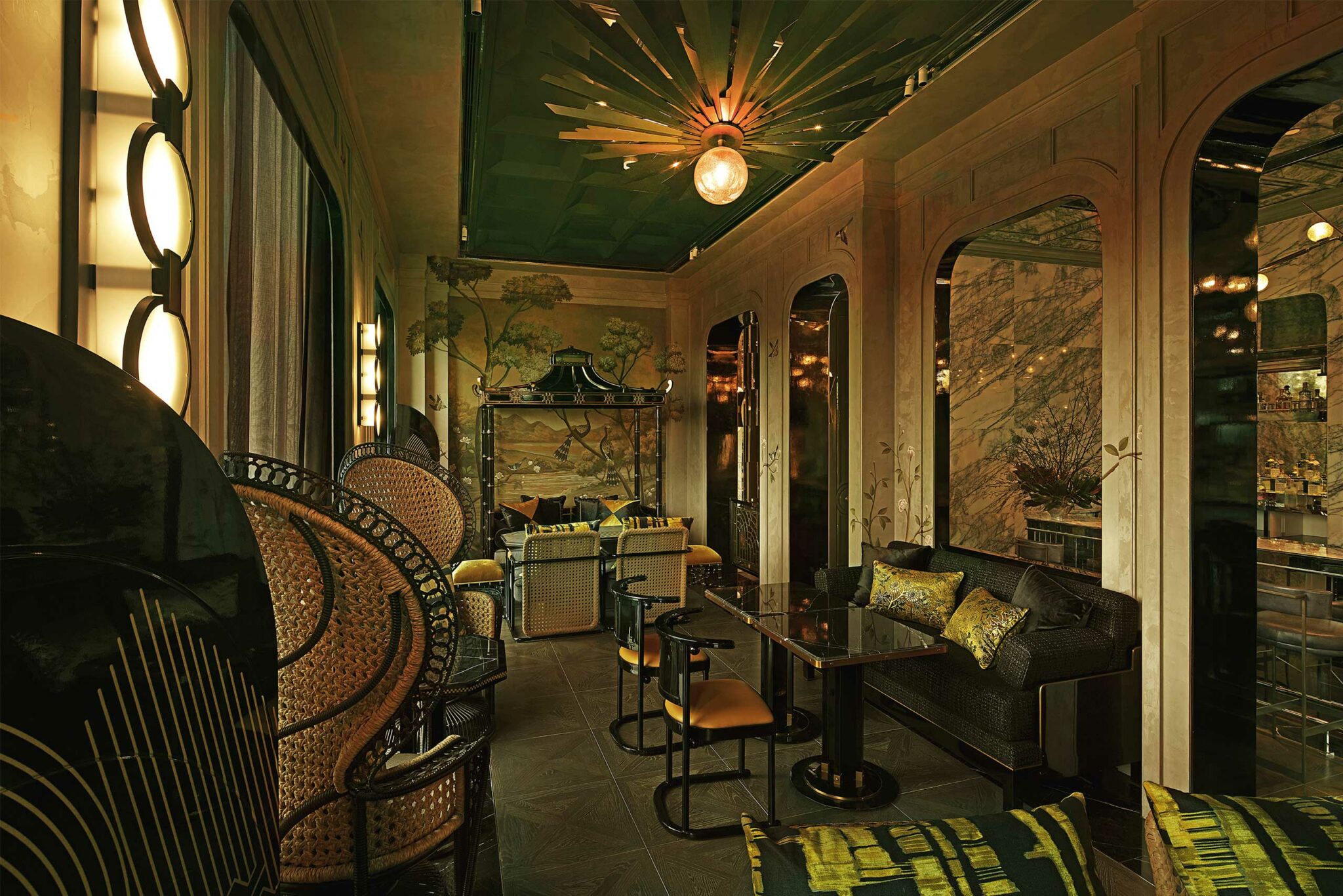Elegant cocktail salon with vintage decor, featuring art deco chairs, a green ceiling with a central light fixture, mirrored walls, and mural paintings.