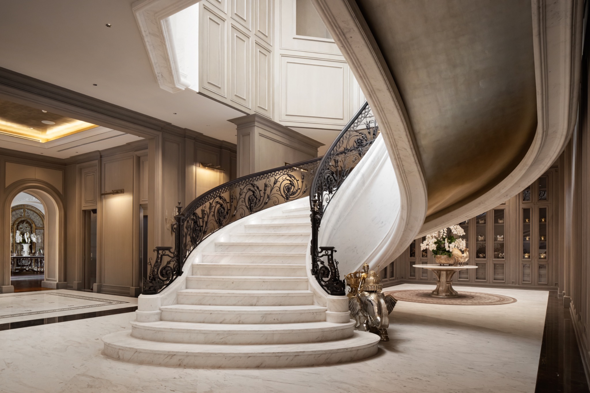 Luxurious marble staircase with ornate iron railing ascends in a grand foyer. Elegant decor and soft lighting create an opulent, sophisticated atmosphere.