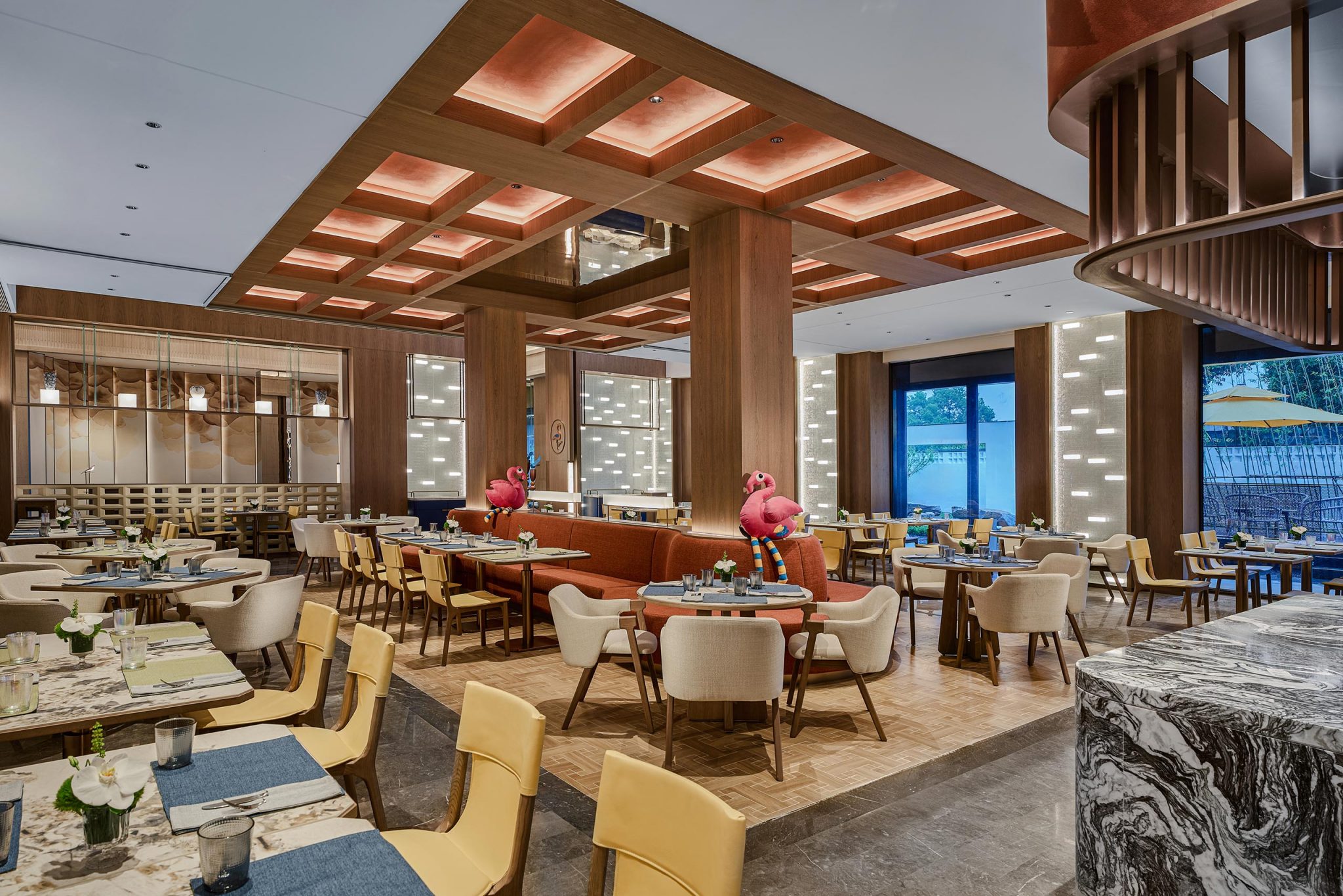 Modern restaurant interior with a warm ambiance, featuring wooden accents, pink flamingo decor, and elegant seating. Large windows offer natural light.