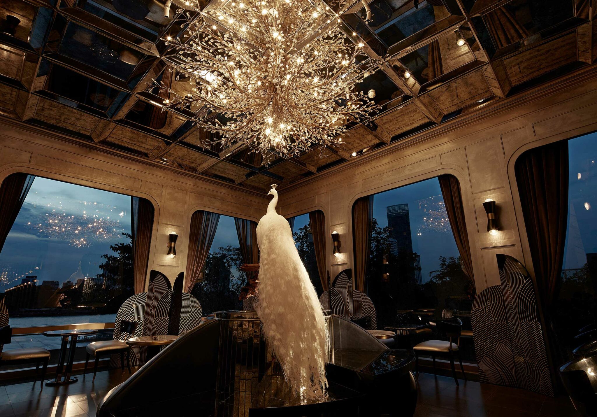 Elegant cocktail salon with a large, crystal chandelier resembling a tree, a white peacock sculpture beneath it. Evening city view through tall windows. Luxurious atmosphere.
