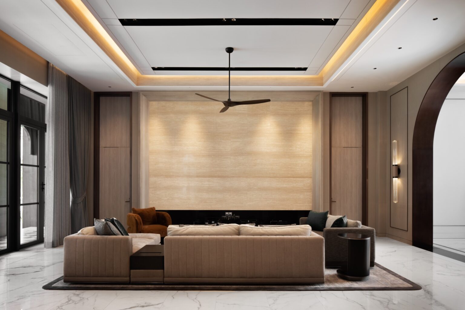 Modern living room with a beige sofa set, brown armchair, and black side table. Features a ceiling fan, large stone wall, and ambient lighting, creating a cozy, elegant atmosphere.