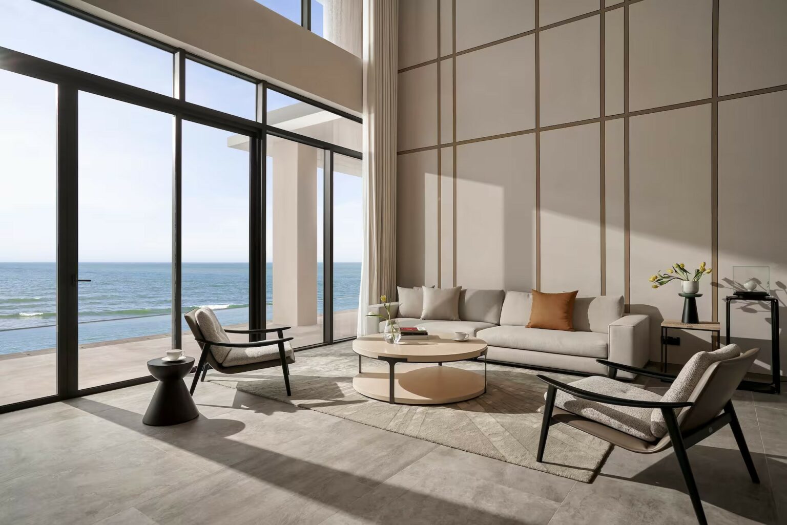 Spacious hotel lounge with large windows overlooking the ocean. Features a neutral-colored sofa, armchairs, round table, and coastal decor.