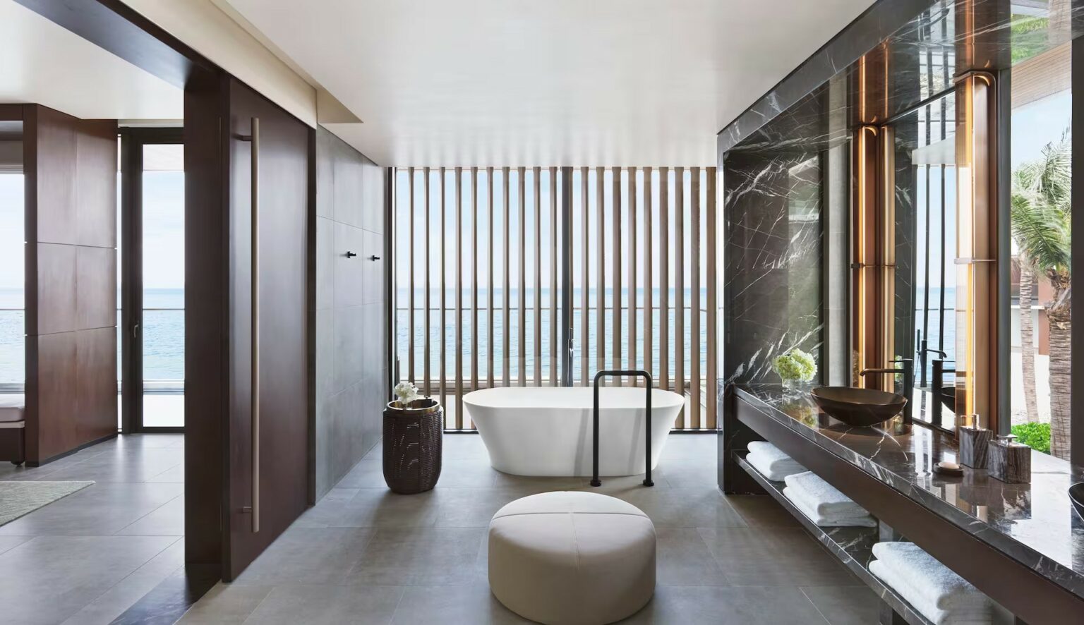 Luxurious hotel bathroom with ocean view, featuring a freestanding bathtub near wooden slats, dark marble sink, and modern decor, exuding a serene, elegant vibe.