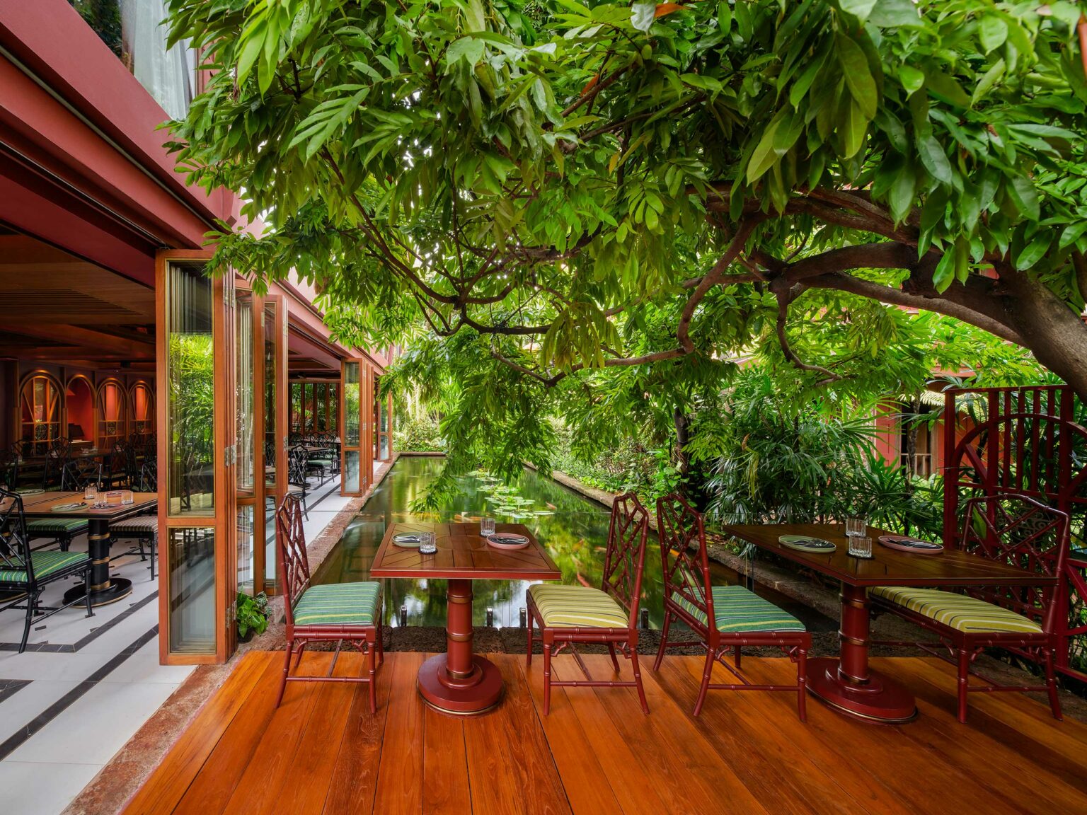 Outdoor restaurant seating under lush trees with wooden tables beside a serene pond. Open sliding doors connect to indoor dining, creating a tranquil vibe.