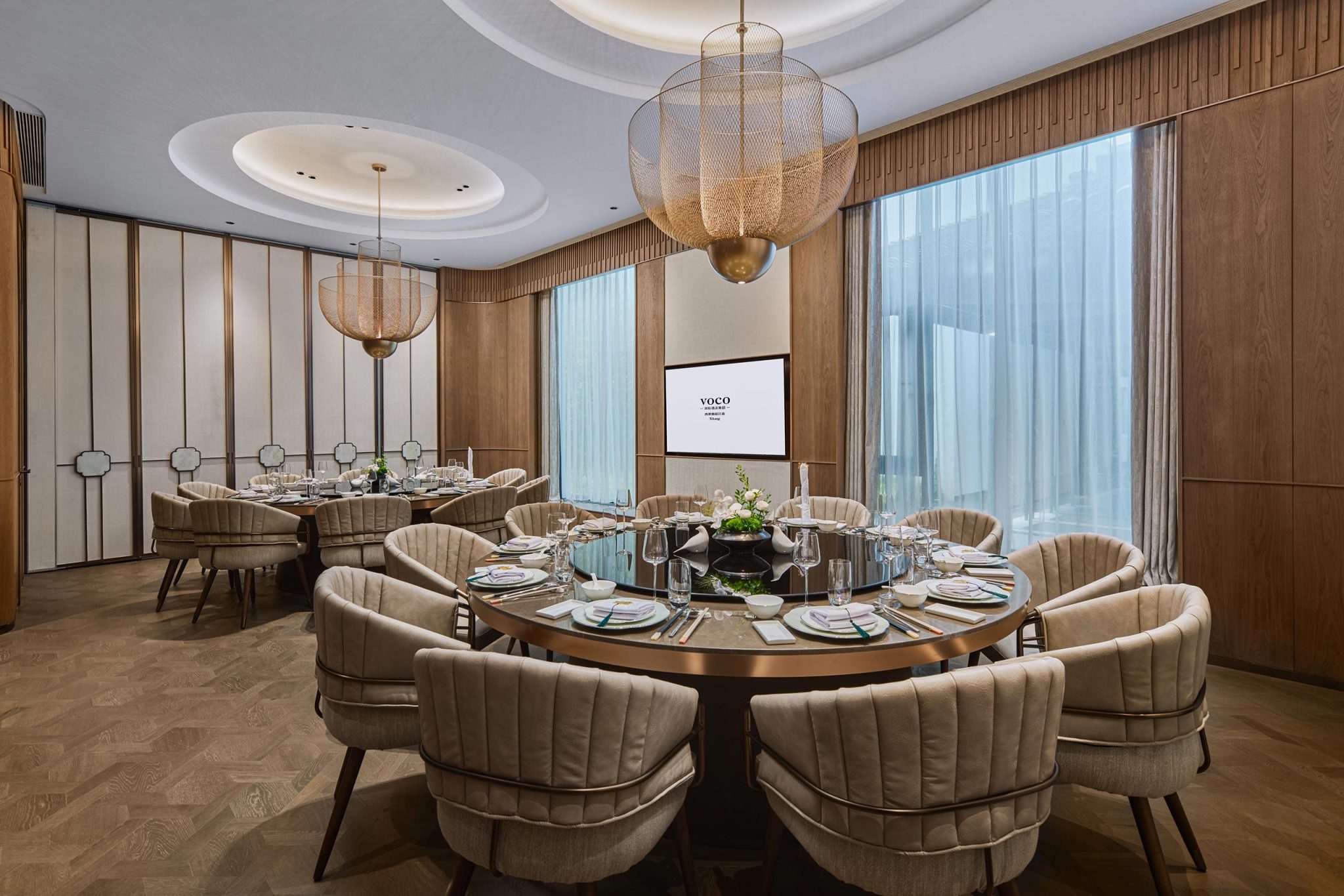 Elegant dining room with a round table set for a formal meal. Neutral-toned chairs surround the table, under large modern chandeliers. Tall windows with sheer curtains add natural light.