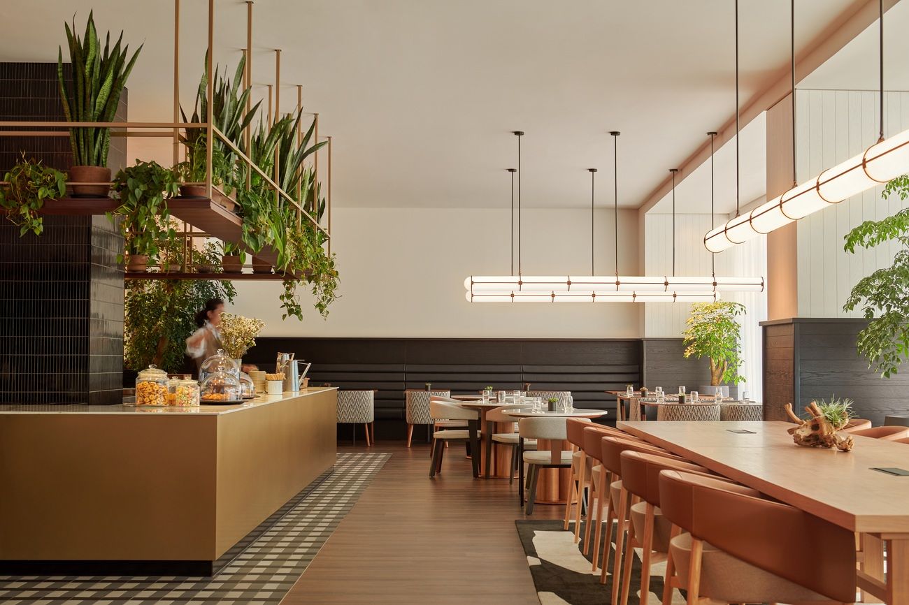 Modern hotel restaurant interior with a warm, inviting atmosphere. Suspended plants and sleek lighting hang over wooden tables and beige chairs. A person stands at a counter.