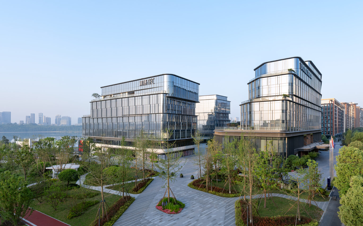 Modern commercial buildings with glass facades and rooftop gardens are surrounded by neatly landscaped greenery. The sky is clear and blue, creating a serene atmosphere.