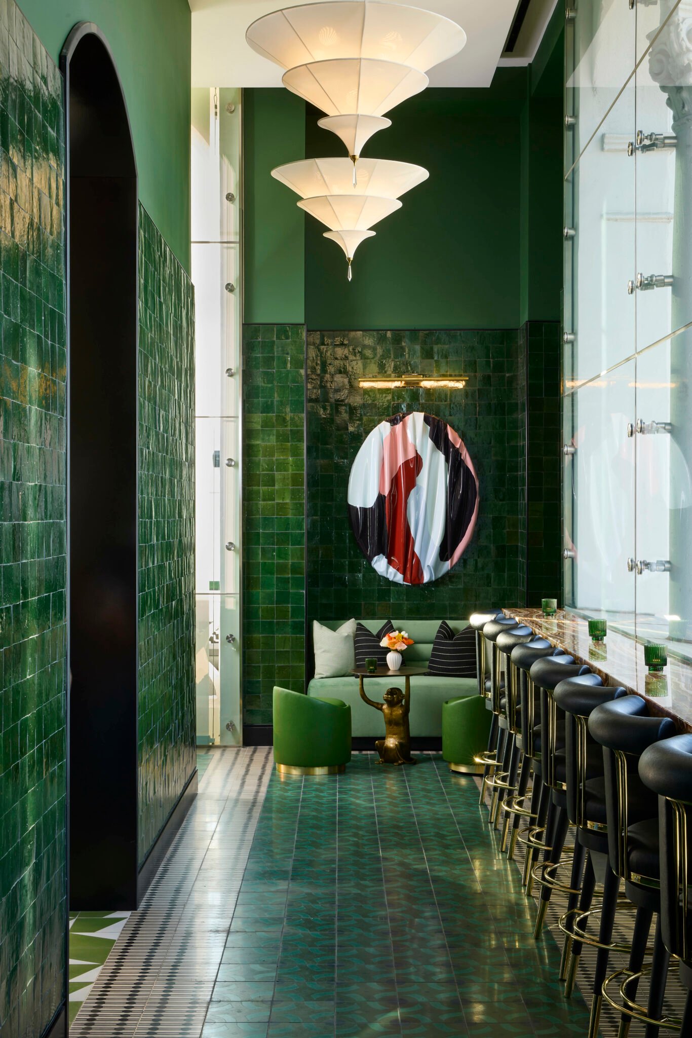 Stylish green-tiled bar with black stools and a round abstract art piece. Soft lighting, cozy seating, and decorative pendant lights create a chic ambiance.