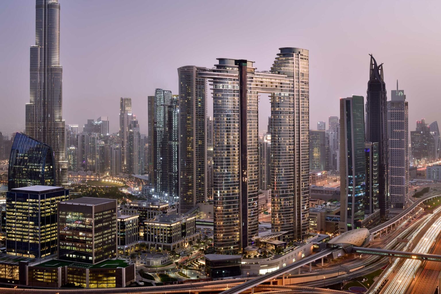 A cityscape at dusk with modern skyscrapers, including a tall, uniquely connected twin-tower structure. Streets below are busy with light trails.