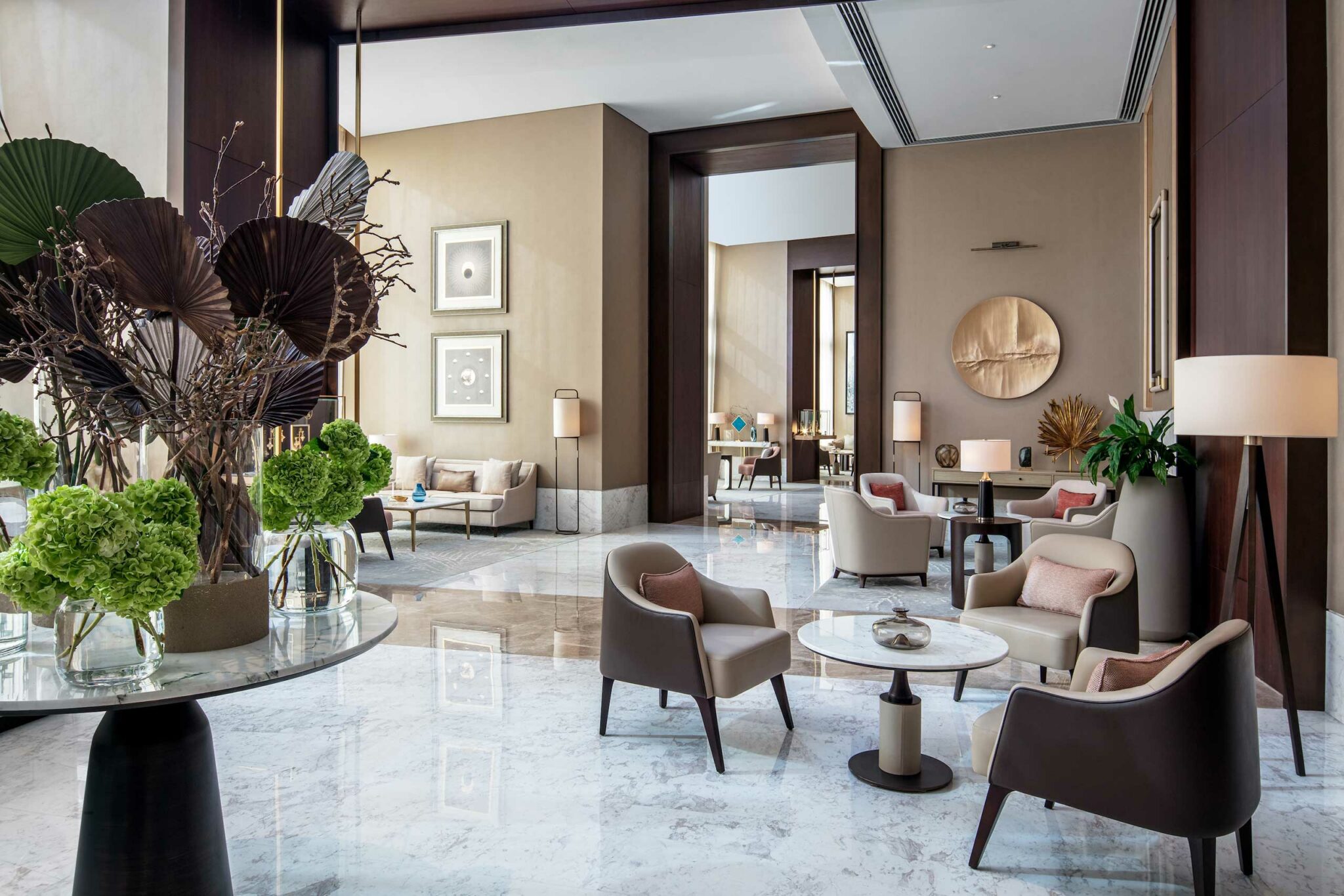 Luxurious hotel lobby with modern beige and brown decor. Elegant chairs and tables, lush green plants, and abstract art create a warm, inviting atmosphere.