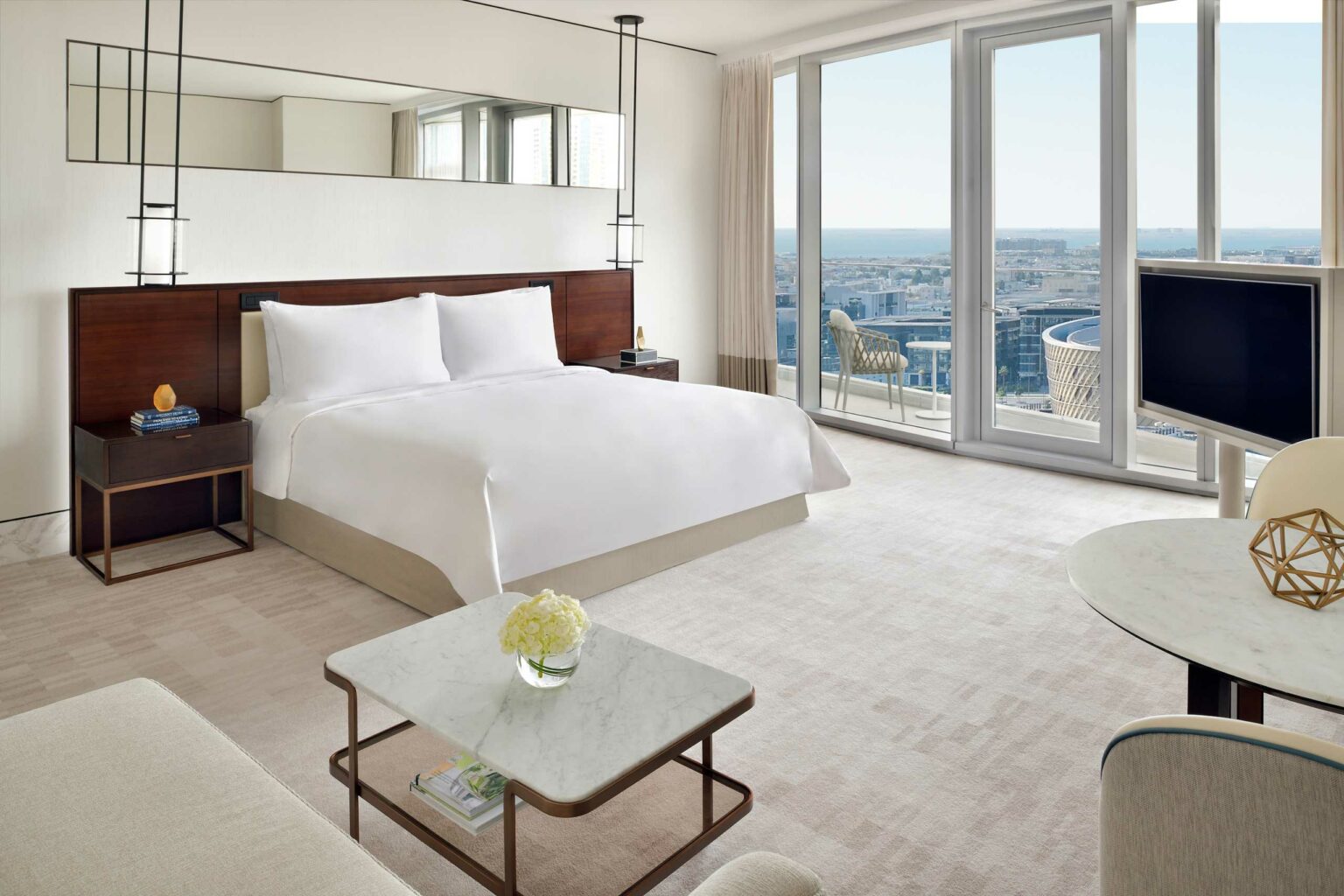 Modern hotel room with a large bed, white linens, and a dark wooden headboard. Floor-to-ceiling windows offer a city skyline view. Elegant and serene atmosphere.