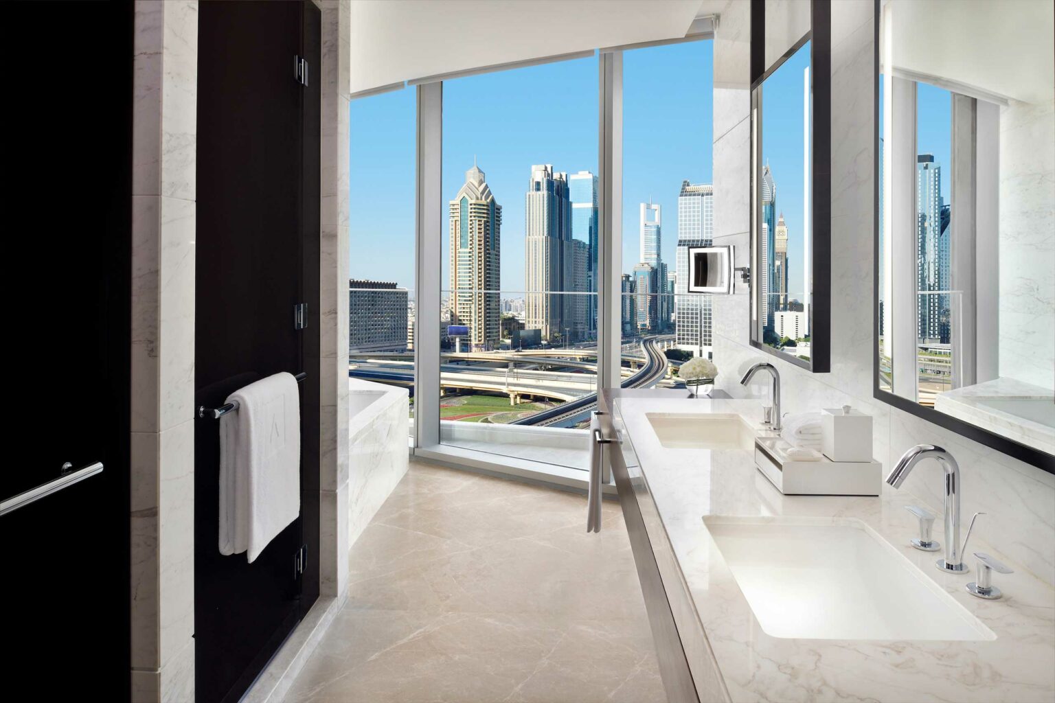 Luxurious suite bathroom with marble countertops and dual sinks, large windows revealing a cityscape of modern skyscrapers under a clear blue sky.