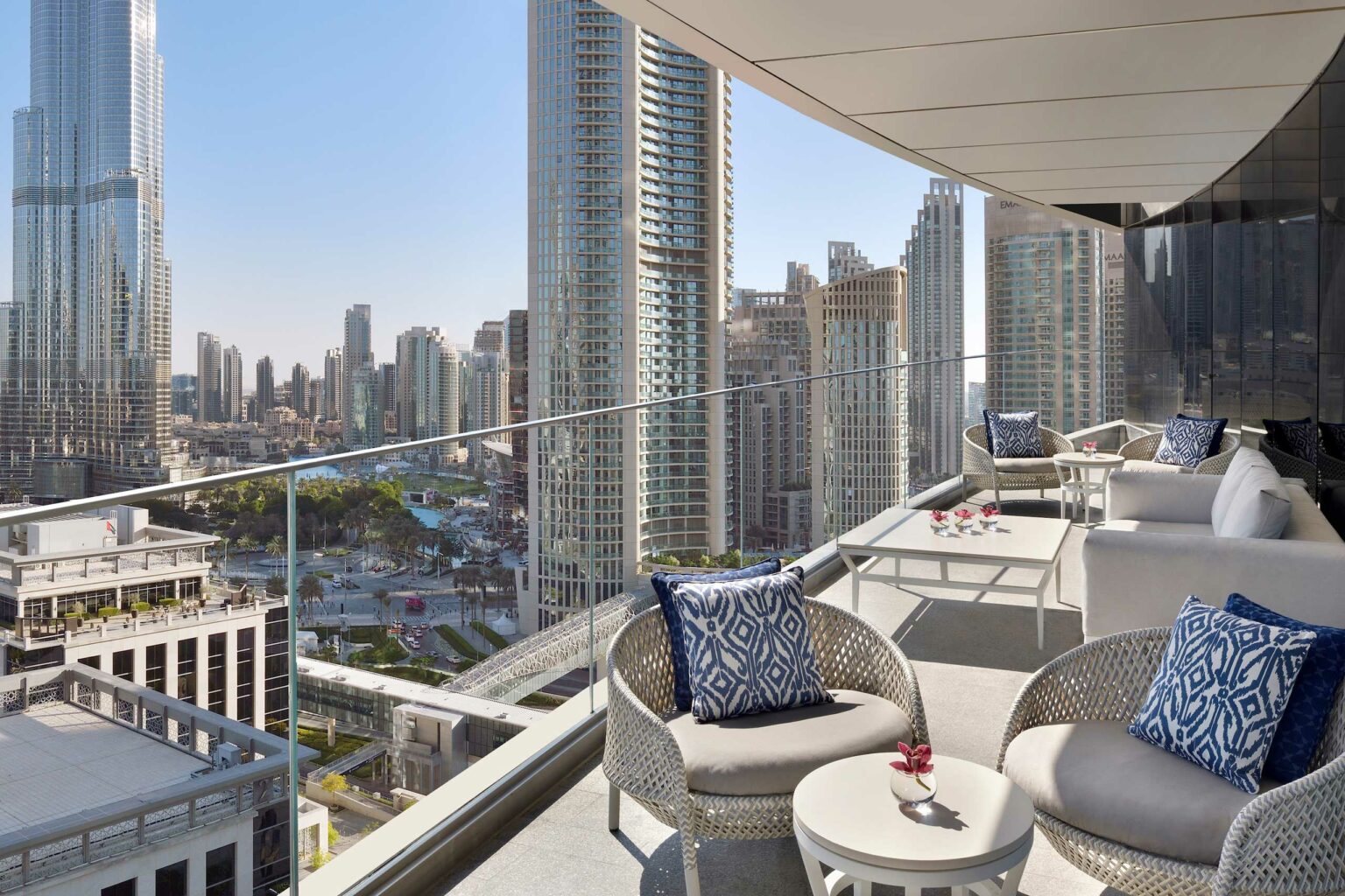 Luxurious hotel balcony overlooking a cityscape with tall skyscrapers, including a distinctive tower. Cozy seating with patterned cushions, exuding a tranquil urban vibe.