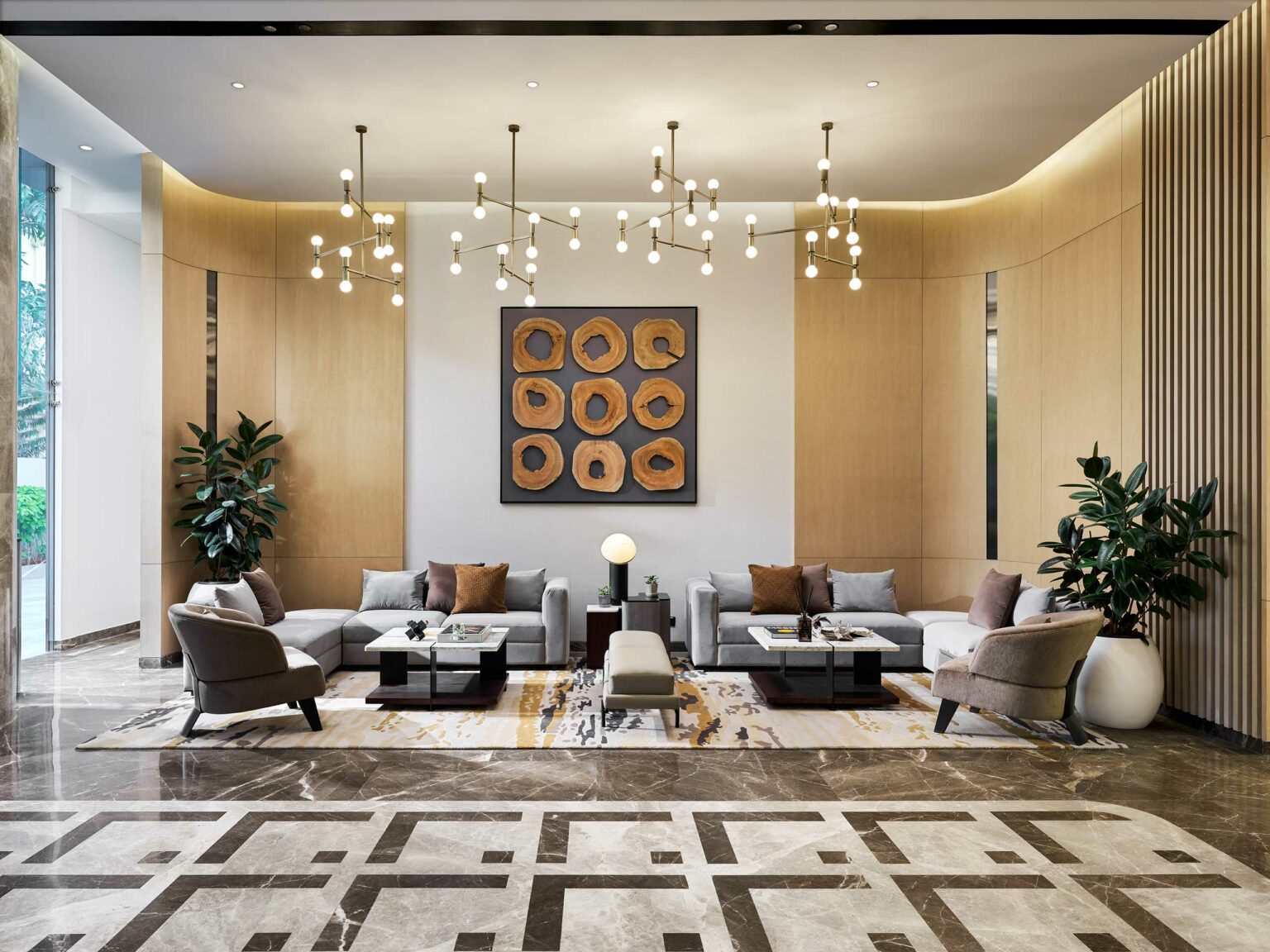 Spacious modern office lounge with gray sofas, geometric wall art, and patterned rugs. Chic chandeliers hang above, with tall green plants adding freshness.