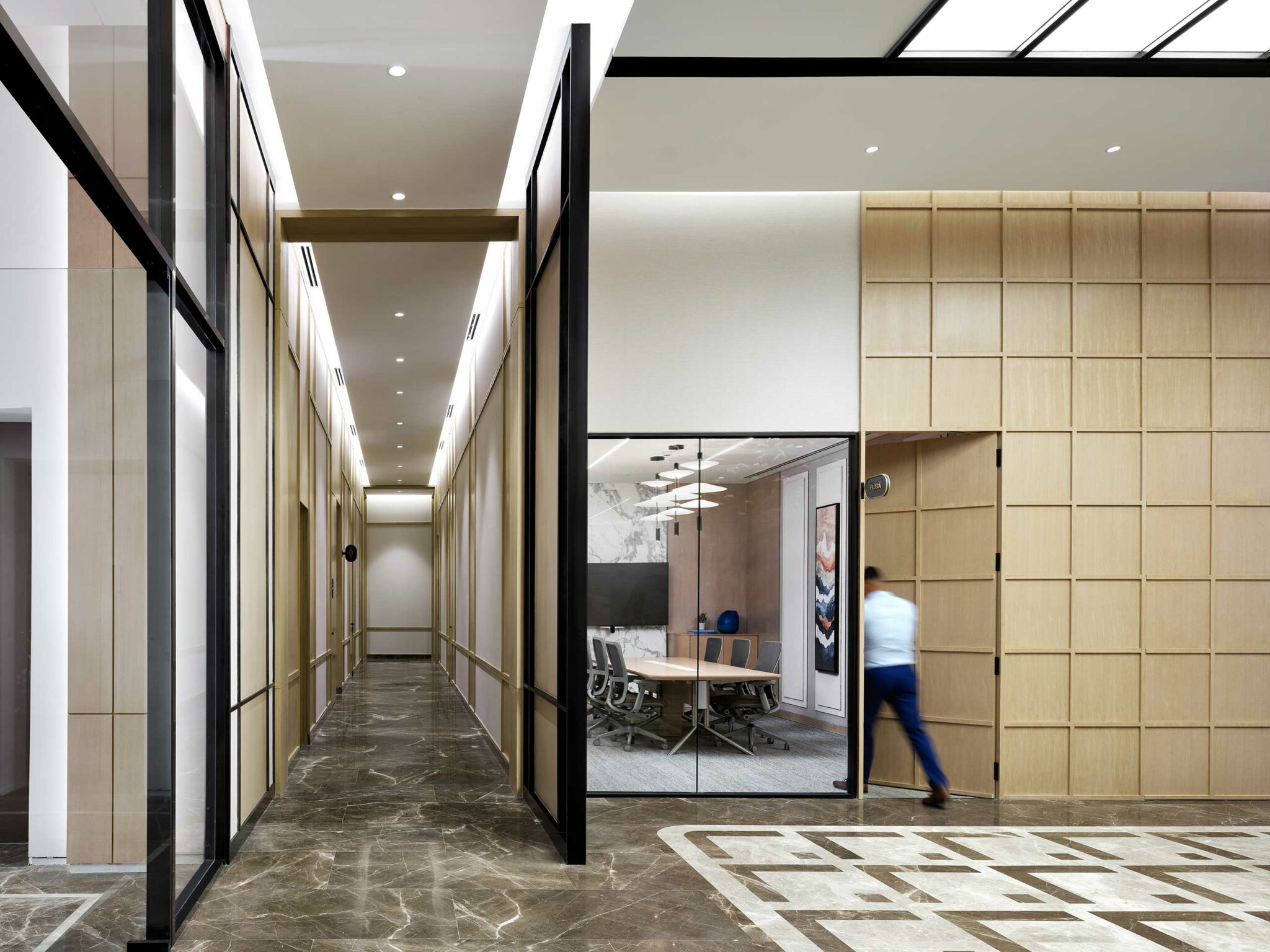 A modern office corridor with a marble floor, leading to a glass-walled conference room. Neutral tones, sleek decor, and a person exiting convey professionalism.
