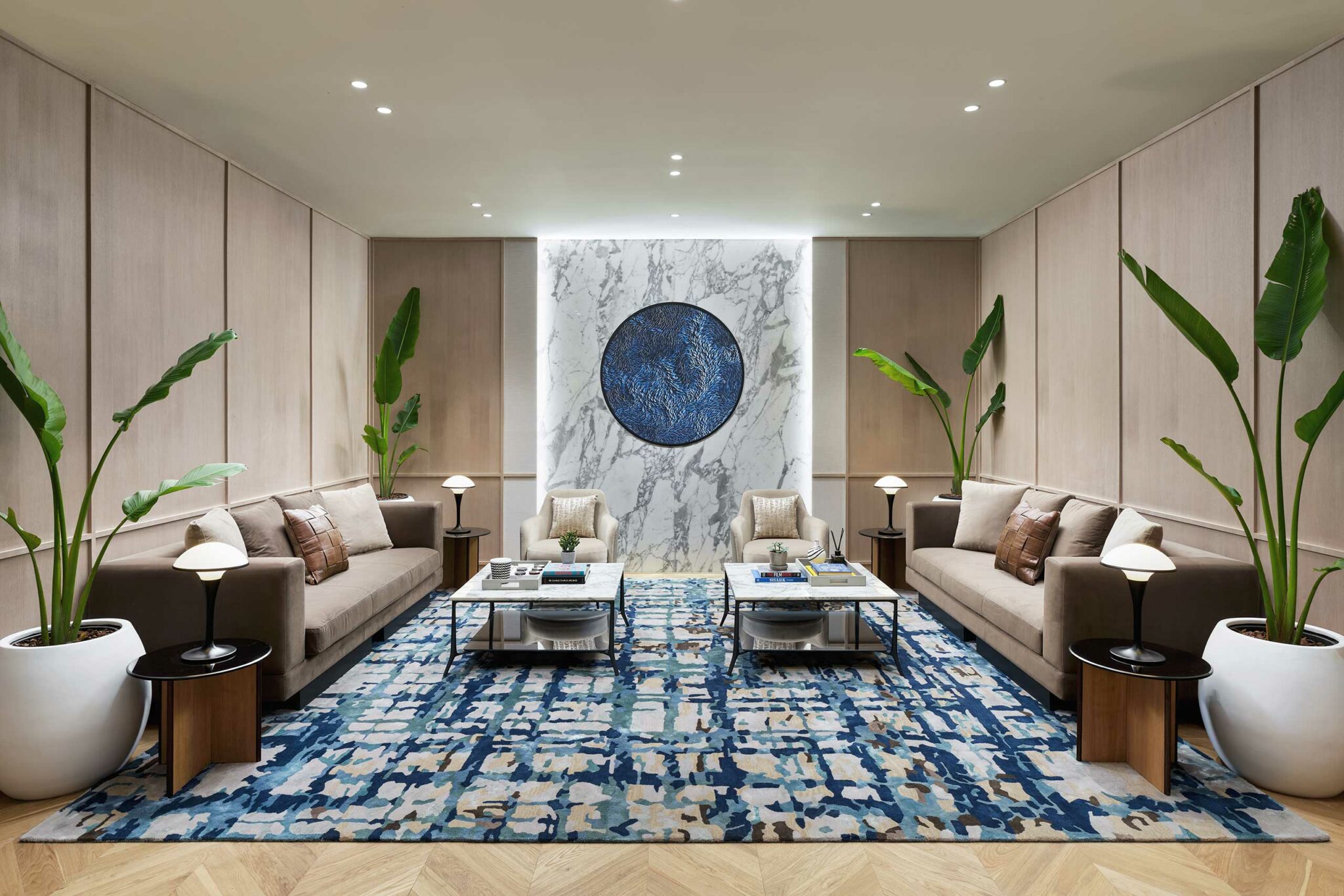 Sophisticated modern office lounge with two beige sofas facing each other, a blue and white rug, marble wall, circular blue artwork, and potted plants.