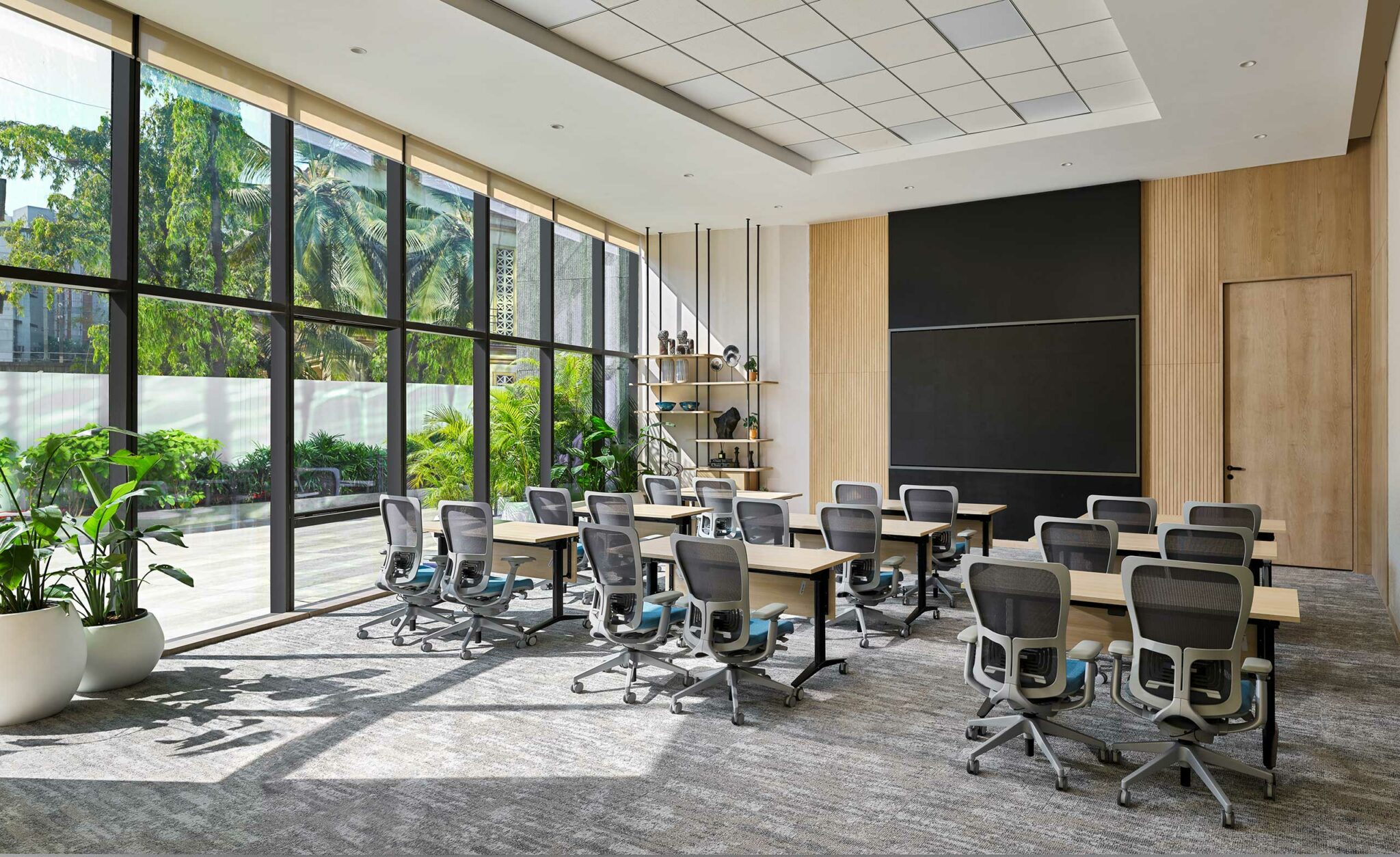 Modern office learning room with large windows revealing greenery outside. Rows of desks with ergonomic chairs face a blank chalkboard, creating a bright, focused atmosphere.