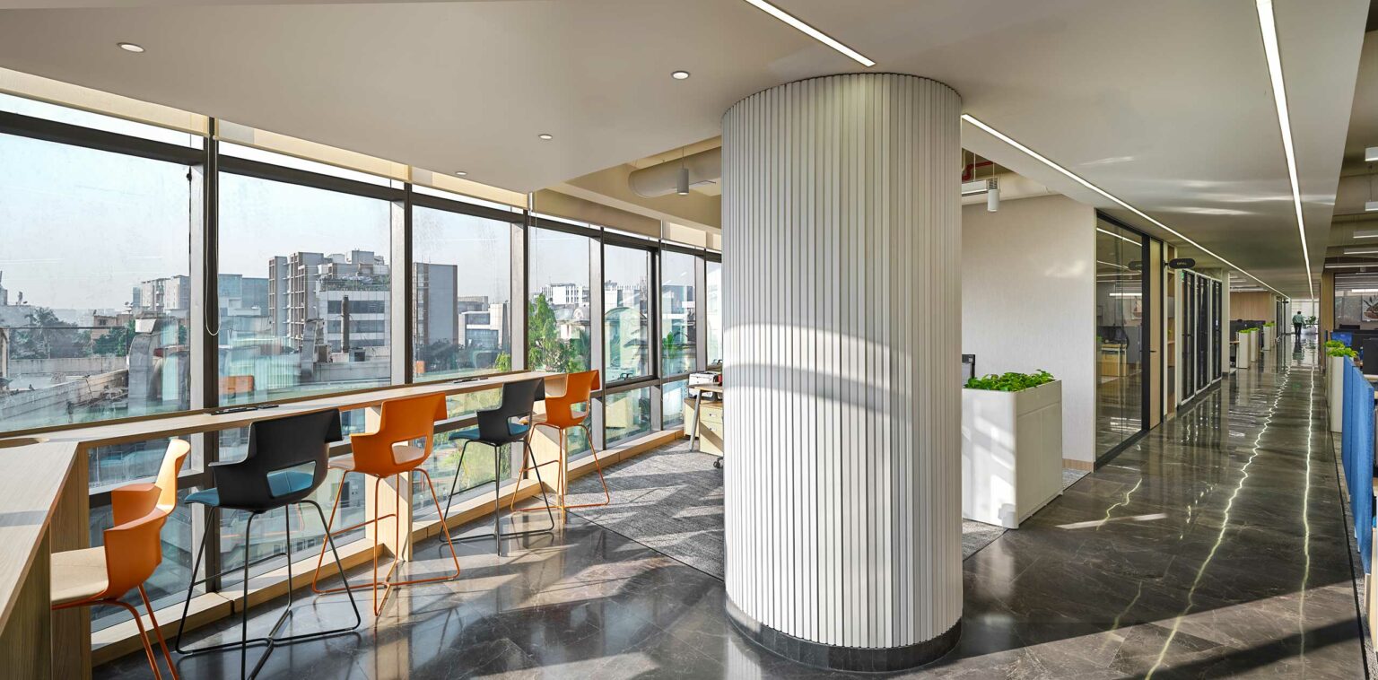 Modern office space with large windows on the left, bright city views, colorful chairs, a round column, and a sleek, polished floor. Airy and open atmosphere.