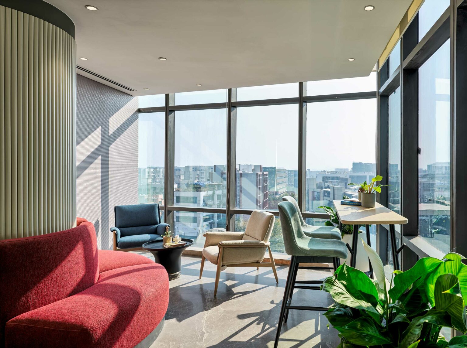 Bright, modern office lounge with large windows offering city views. Features colorful seating, plants, and natural light, creating a calm atmosphere.