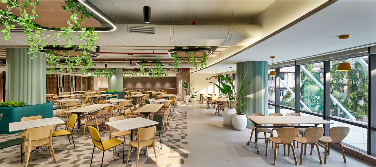 Stylish modern office cafeteria with wooden tables, yellow chairs, and potted plants. Ceiling greenery and large windows create a bright, inviting atmosphere.