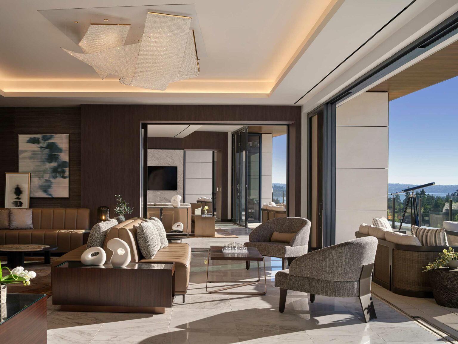 Luxurious hotel lounge with modern beige furniture, plush armchairs, and artistic chandelier. Large windows offer an expansive view of a sunny outdoor terrace.