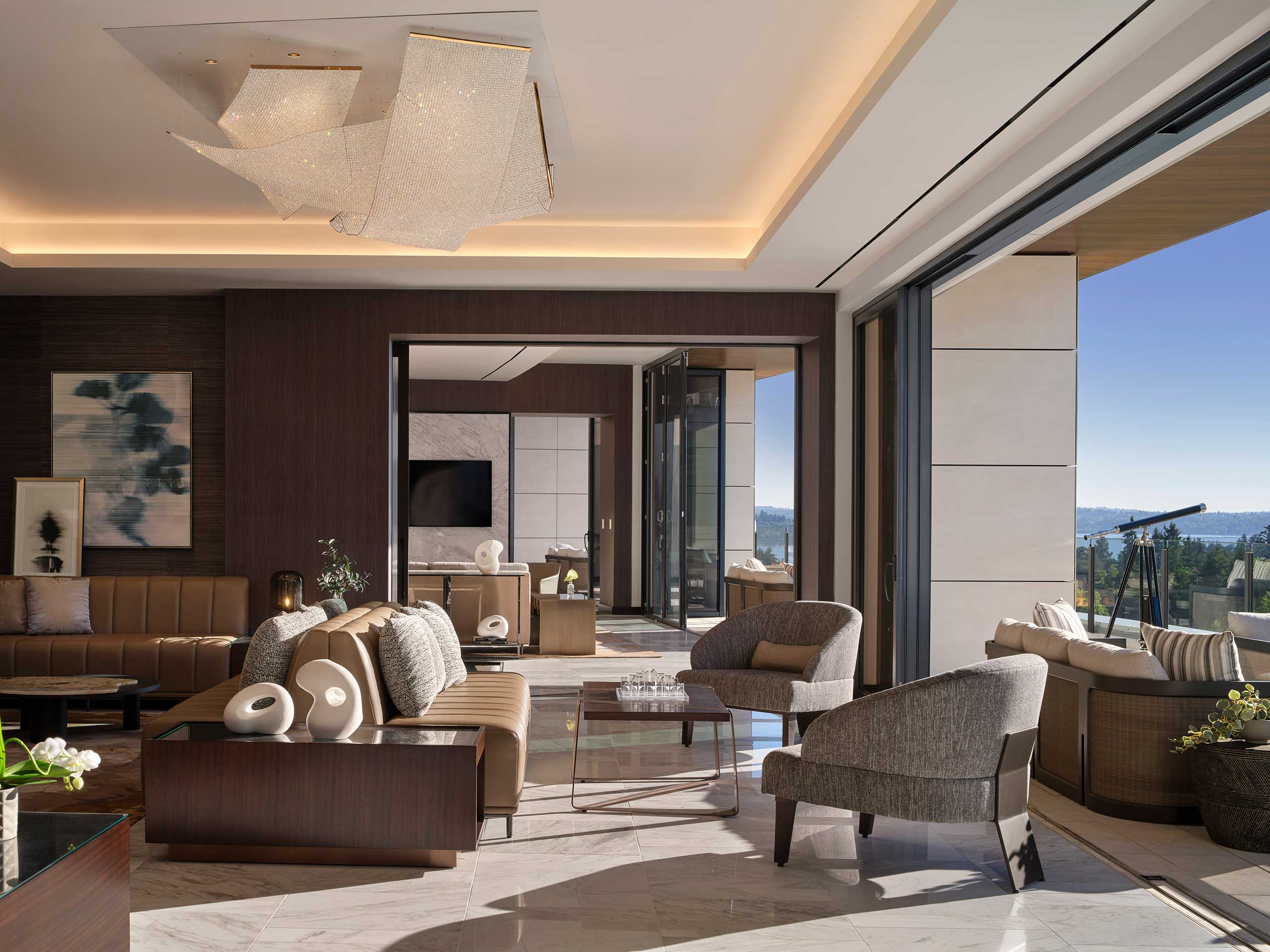 Luxurious hotel lounge with modern beige furniture, plush armchairs, and artistic chandelier. Large windows offer an expansive view of a sunny outdoor terrace.