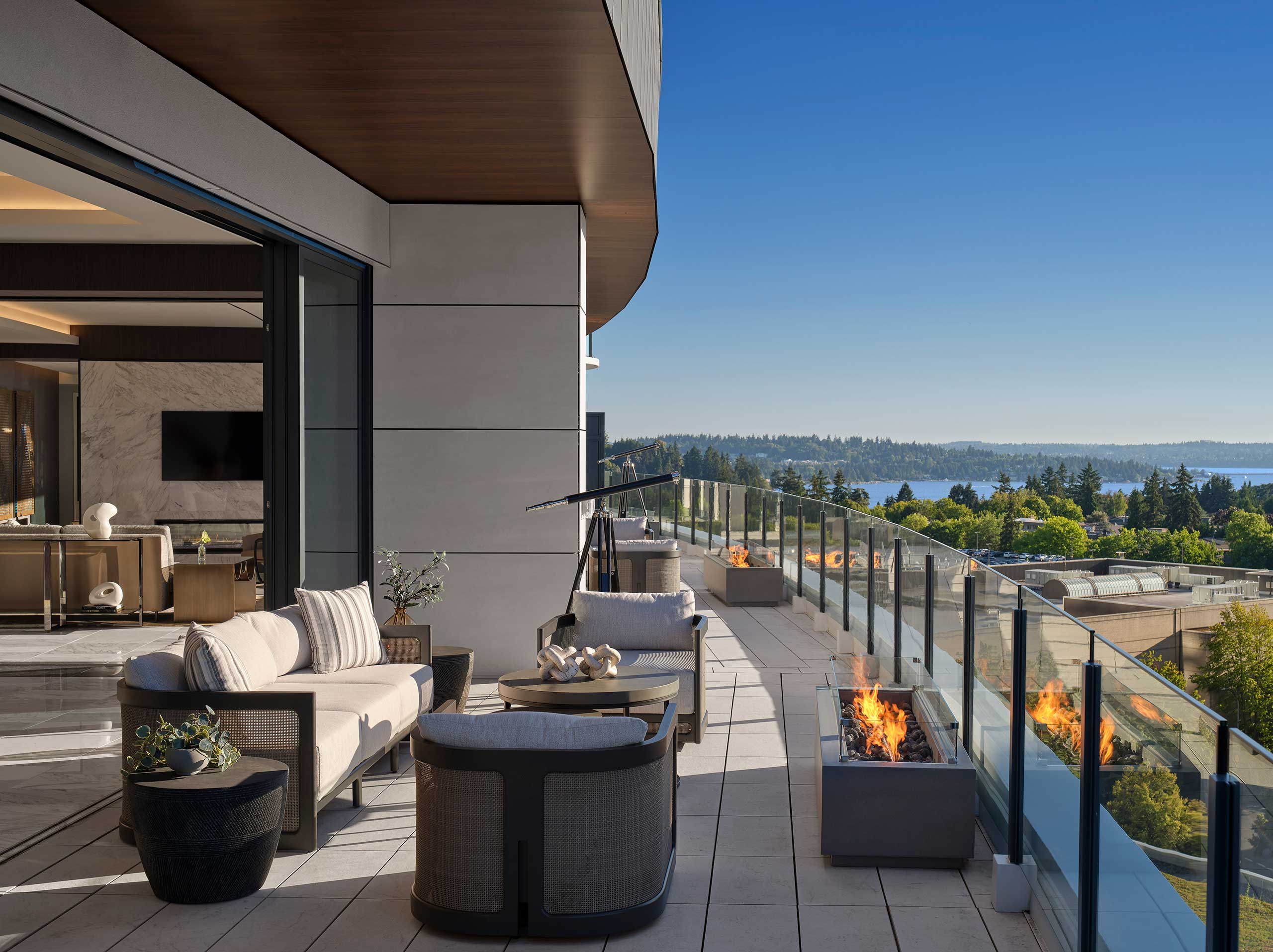 Luxurious hotel rooftop terrace with modern seating, a sleek glass railing, and fire pits. Overlooks lush trees and a serene lake under a clear blue sky.