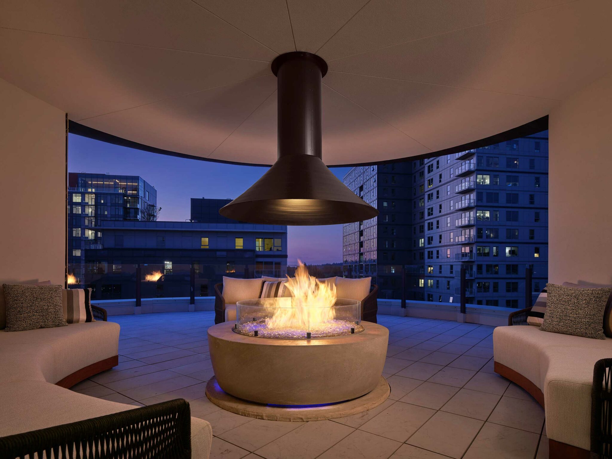 Stylish hotel rooftop patio at dusk with a circular fire pit under a chimney, surrounded by cushioned sofas. Cityscape and modern buildings in the background.