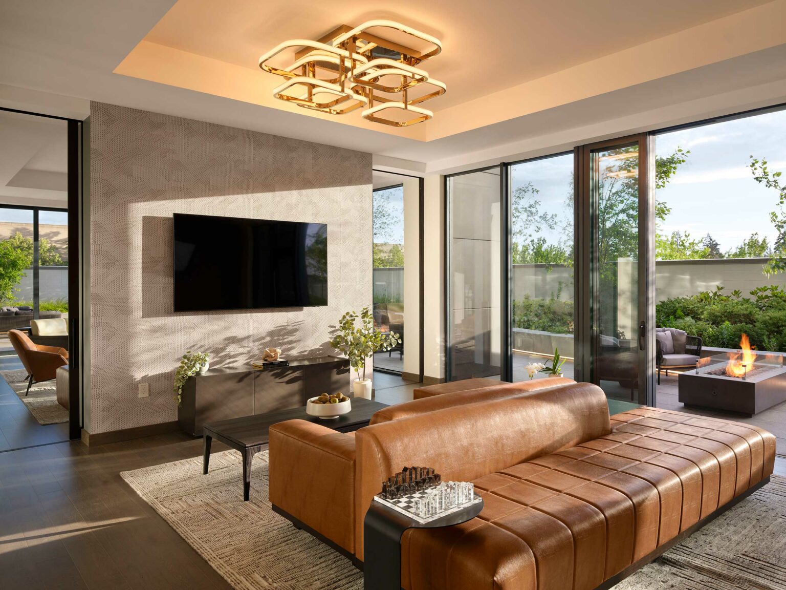 Modern suite living room with a large leather sofa facing a wall-mounted TV, glass doors opening to a patio with a fire pit. Warm, inviting atmosphere.