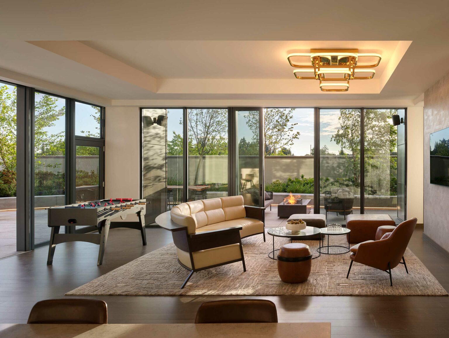 Modern hotel lounge with large windows showcasing a garden view. Includes a beige sofa, armchairs, foosball table, and a ceiling light fixture. Cozy and inviting.