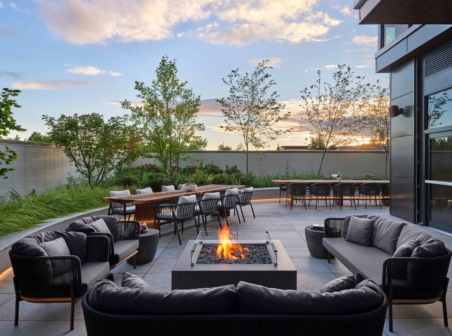 Modern hotel outdoor patio with fire pit, gray sofas, and dining tables. Surrounded by greenery and sunset sky, creating a cozy, inviting atmosphere.