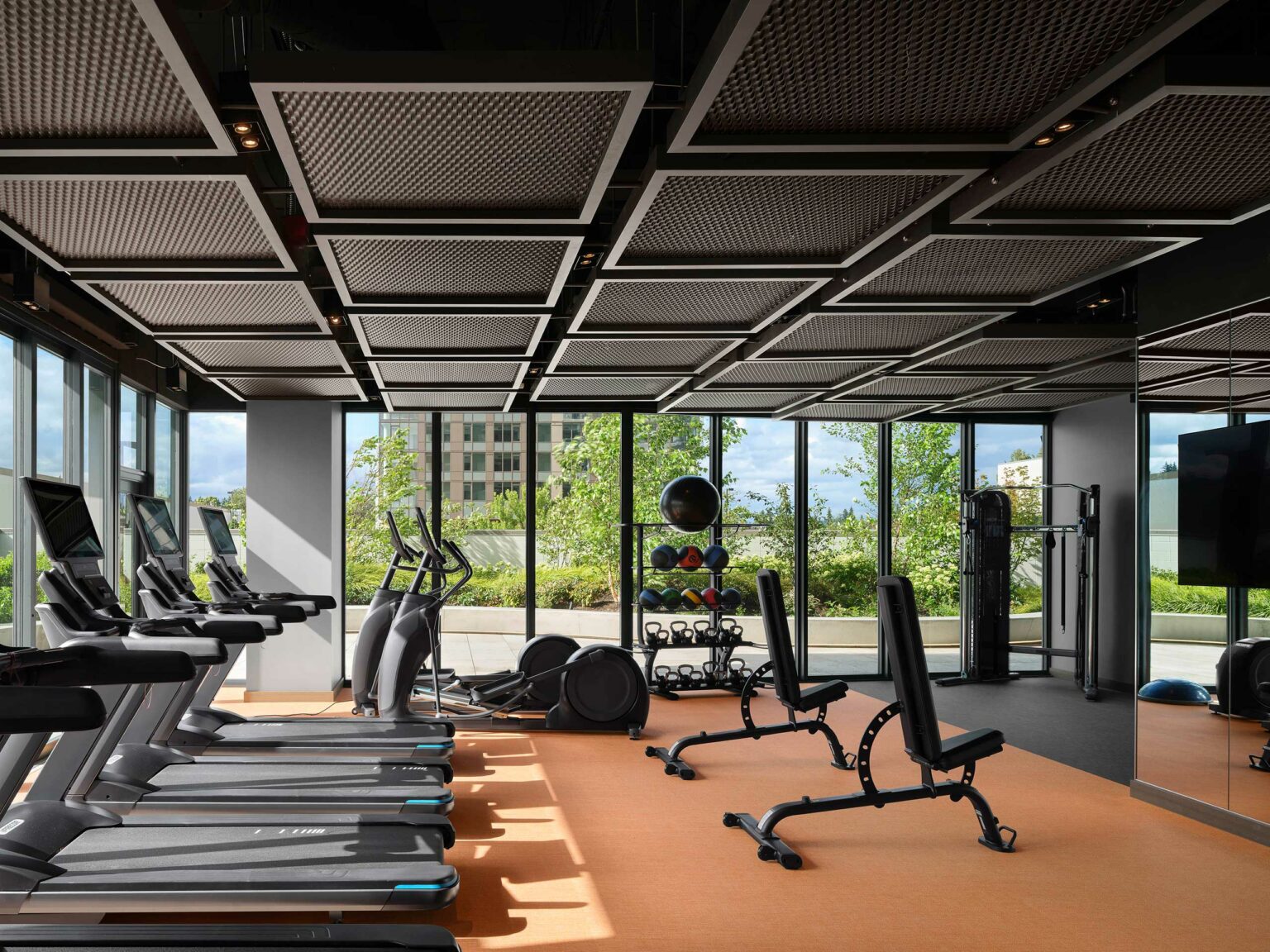 Modern hotel gym with treadmills, benches, and weights on an orange floor. Large windows reveal a view of green trees and a cityscape, creating a bright and inviting atmosphere.
