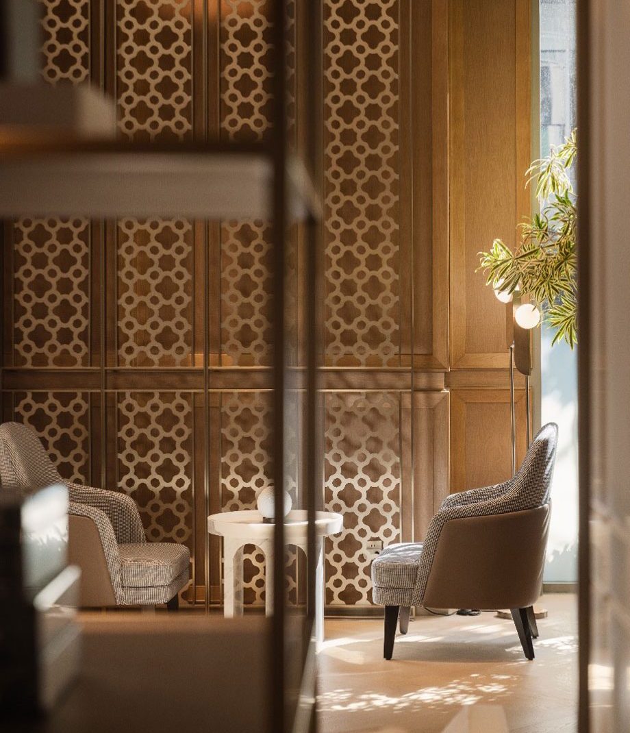 Cozy hotel seating area with two gray armchairs and a small white table against a decorative wooden lattice wall. A floor lamp and plant add warmth.