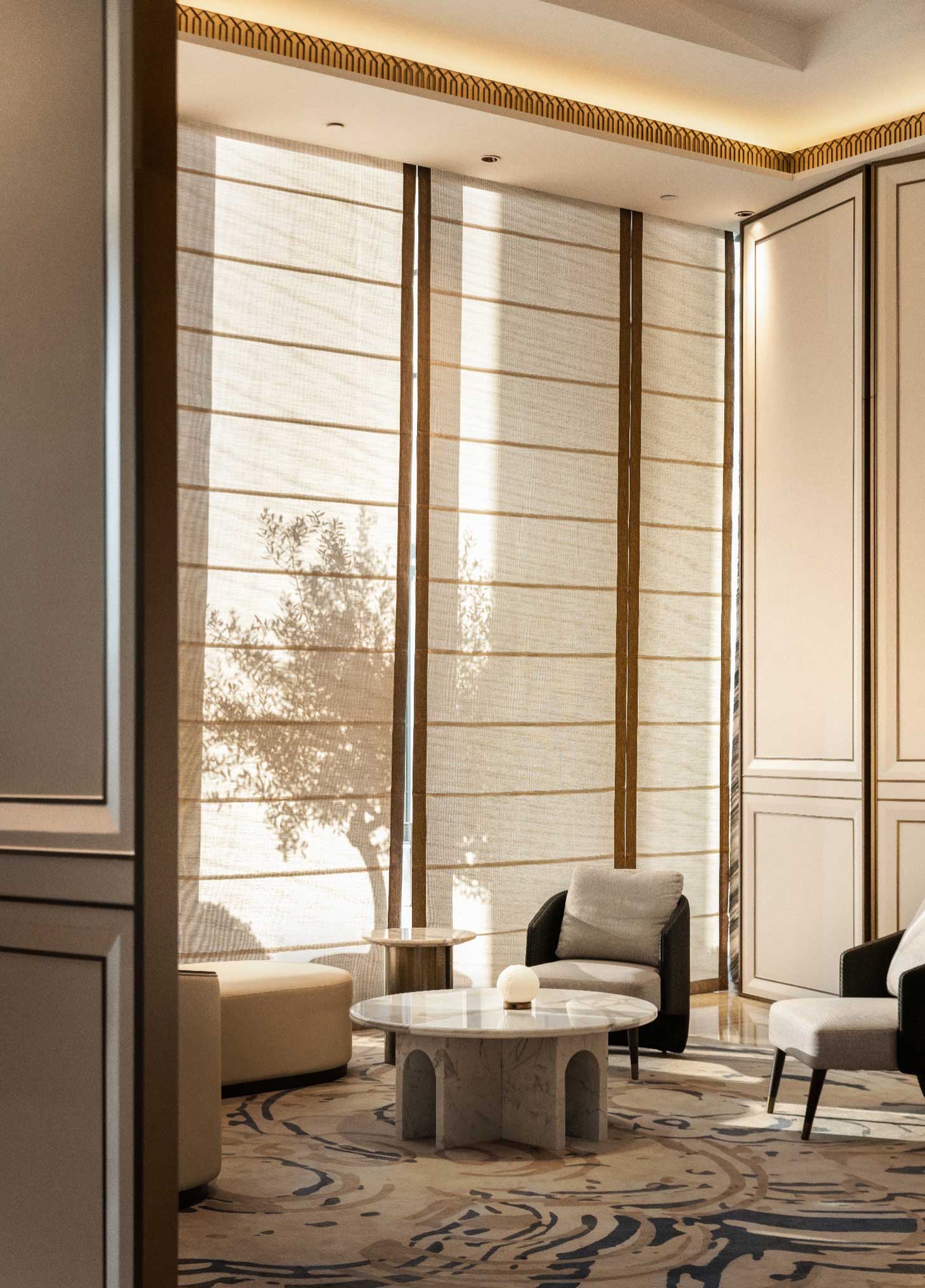 Sophisticated hotel lounge with large, sheer blinds filtering sunlight, casting a tree shadow. Features modern furniture and a marble table, conveying elegance and calm.