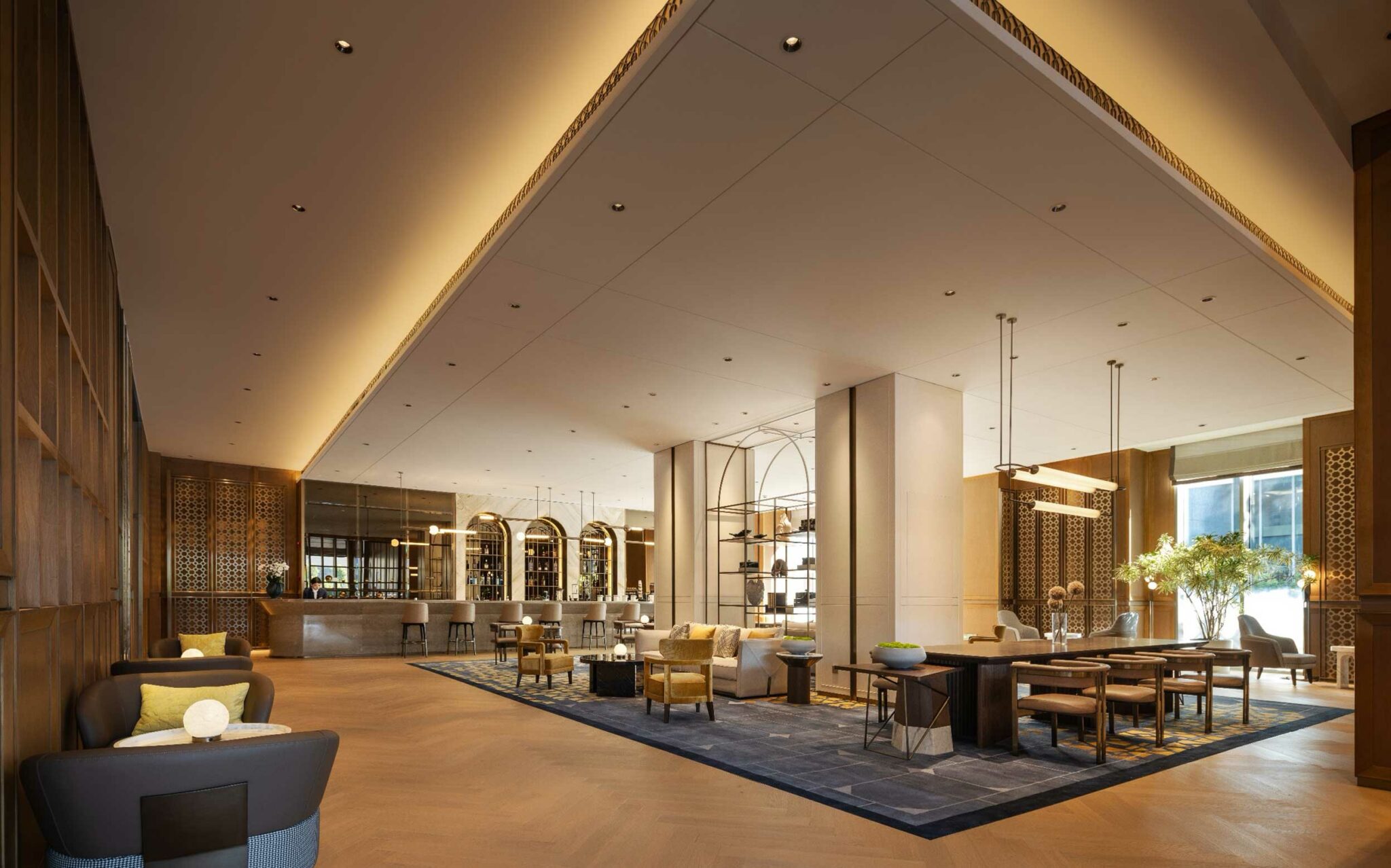 Luxurious hotel lobby with high ceilings and warm lighting. Features plush seating, elegant wooden decor, a central bar, and large windows with natural light.