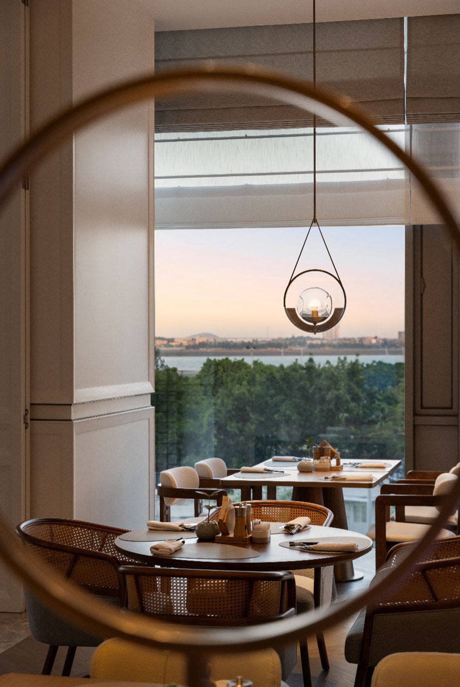 Elegant hotel restaurant with scenic window view, featuring modern circular light fixture. Calm ambiance with distant cityscape and nature at sunset.
