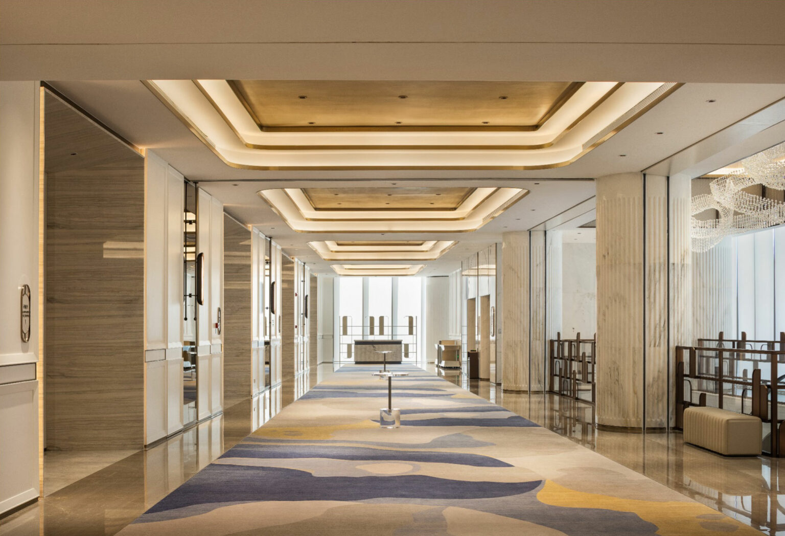 Luxurious hotel hallway with a patterned carpet, elegant ceiling lights, marble columns, and large mirrors, leading to a bright window. Calm and sophisticated ambiance.
