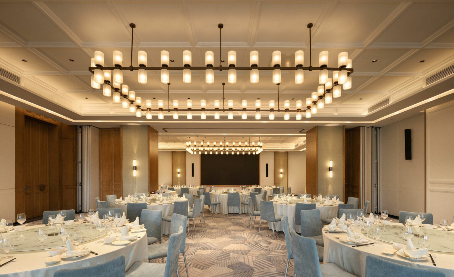 Elegant hotel banquet hall with soft lighting features round tables set for dining, surrounded by light blue chairs. A large chandelier adds a warm, inviting glow.