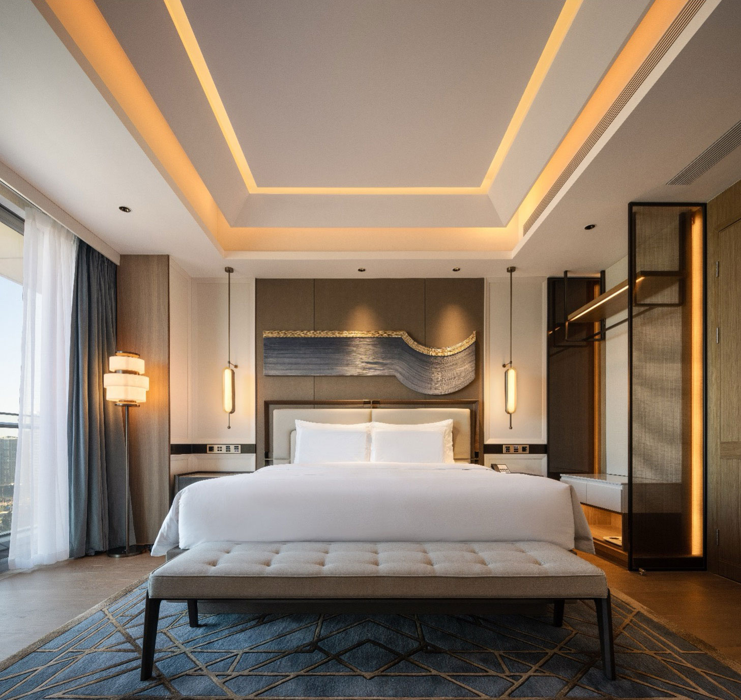 Modern hotel room with a plush white bed, tufted bench, geometric rug, and elegant wall art. Warm lighting from ceiling and side lamps creates a cozy ambiance.