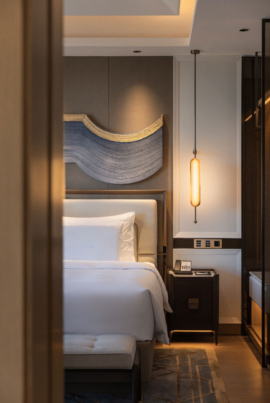 Elegant hotel room interior with a neatly made bed, soft white linens, a modern wooden nightstand, and a sleek, elongated pendant light creating a cozy ambiance.