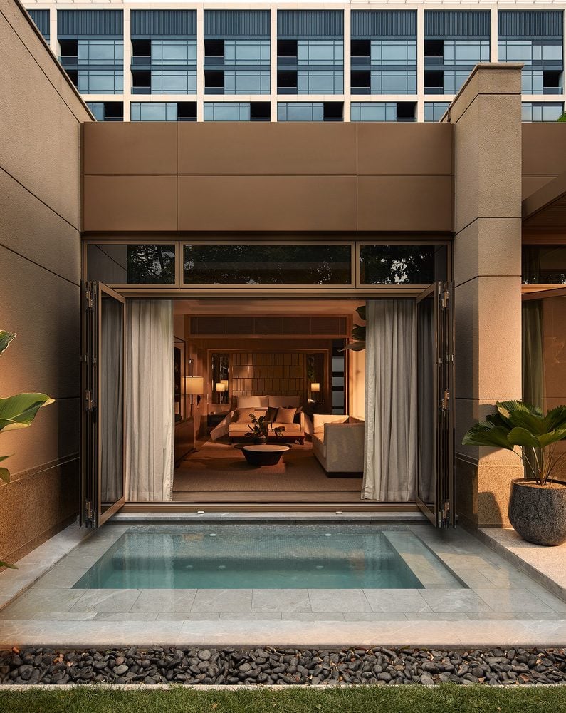 Elegant modern hotel exterior with an open living room visible through large glass doors. A small pool is in the foreground, surrounded by lush plants.
